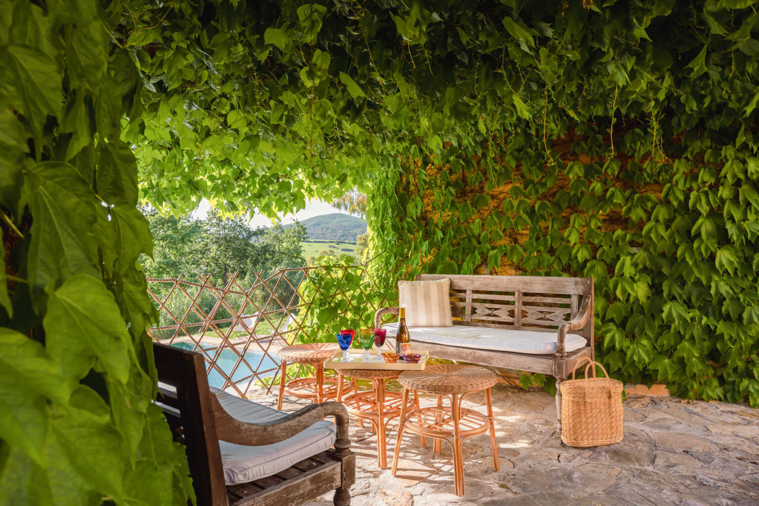 Cozy outdoor seating area surrounded by lush green vines, featuring a wooden bench, two tables, and decorative drinks, ideal for relaxation and enjoying nature. | Only on SopranoVillas