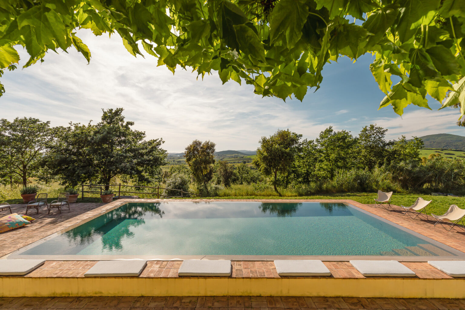 Serene outdoor pool surrounded by lush greenery and scenic hills, featuring comfortable lounge chairs and a clear blue sky, ideal for relaxation and leisure. | Only on SopranoVillas