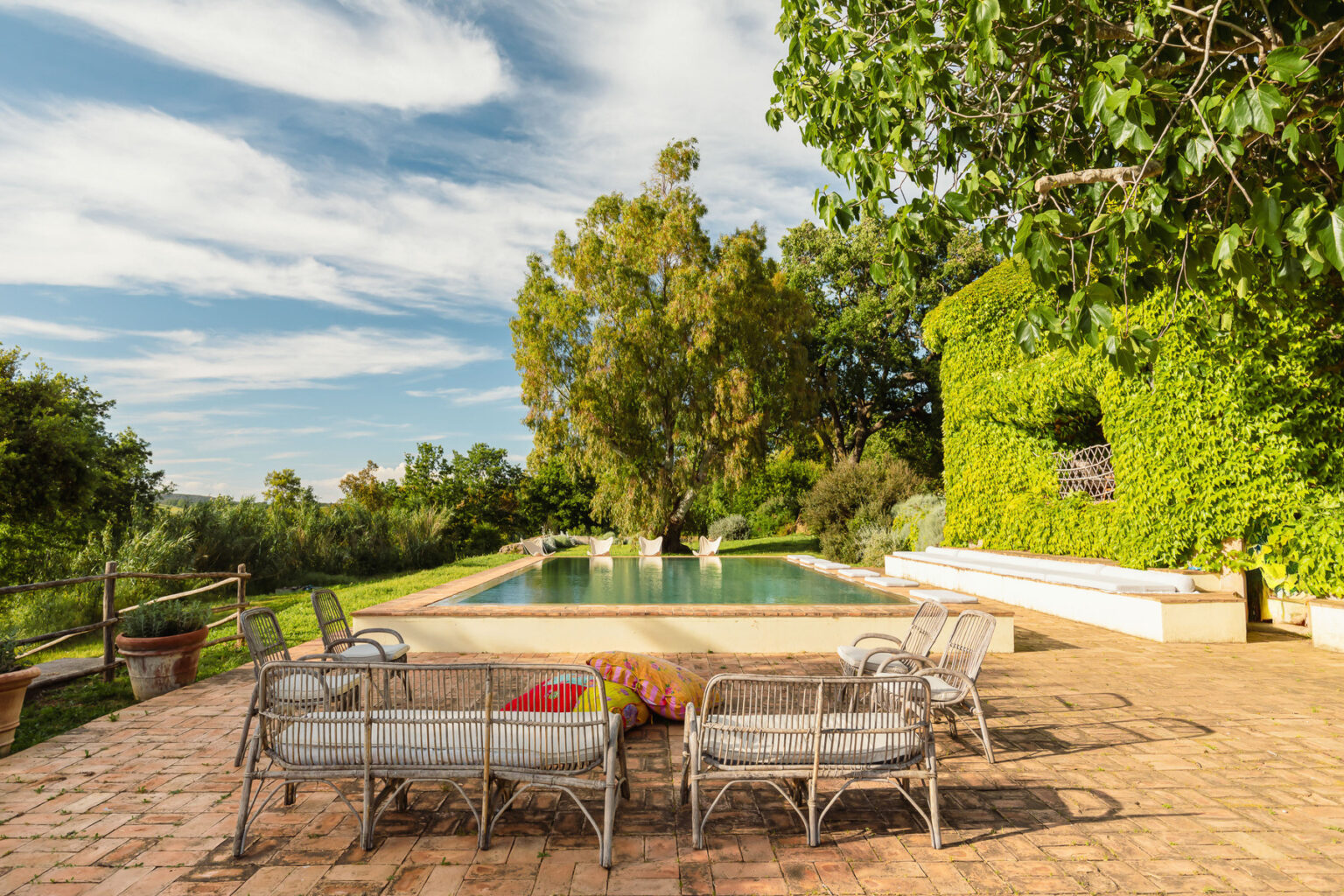 Serene outdoor pool area surrounded by lush greenery, featuring stylish wicker seating, vibrant cushions, and a clear blue sky. Ideal for relaxation and leisure in a tranquil setting. | Only on SopranoVillas