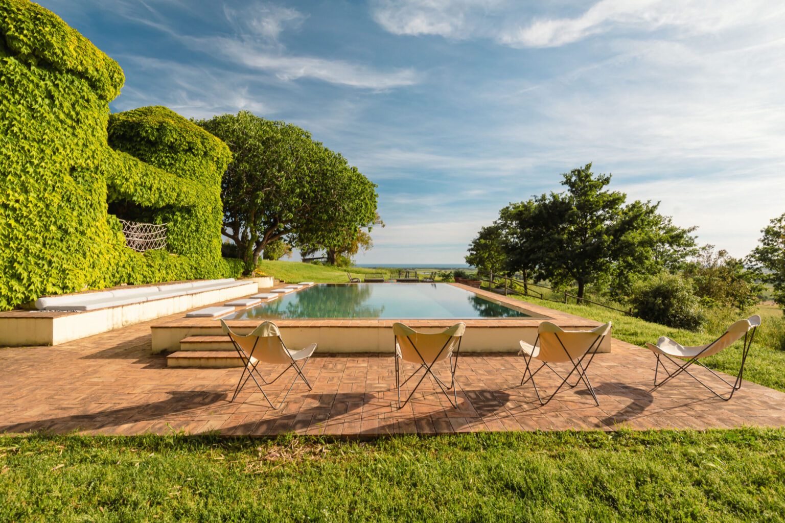 Lush green landscape featuring a serene swimming pool surrounded by modern lounge chairs, with vibrant foliage and a clear blue sky in the background, ideal for relaxation and outdoor gatherings. | Only on SopranoVillas