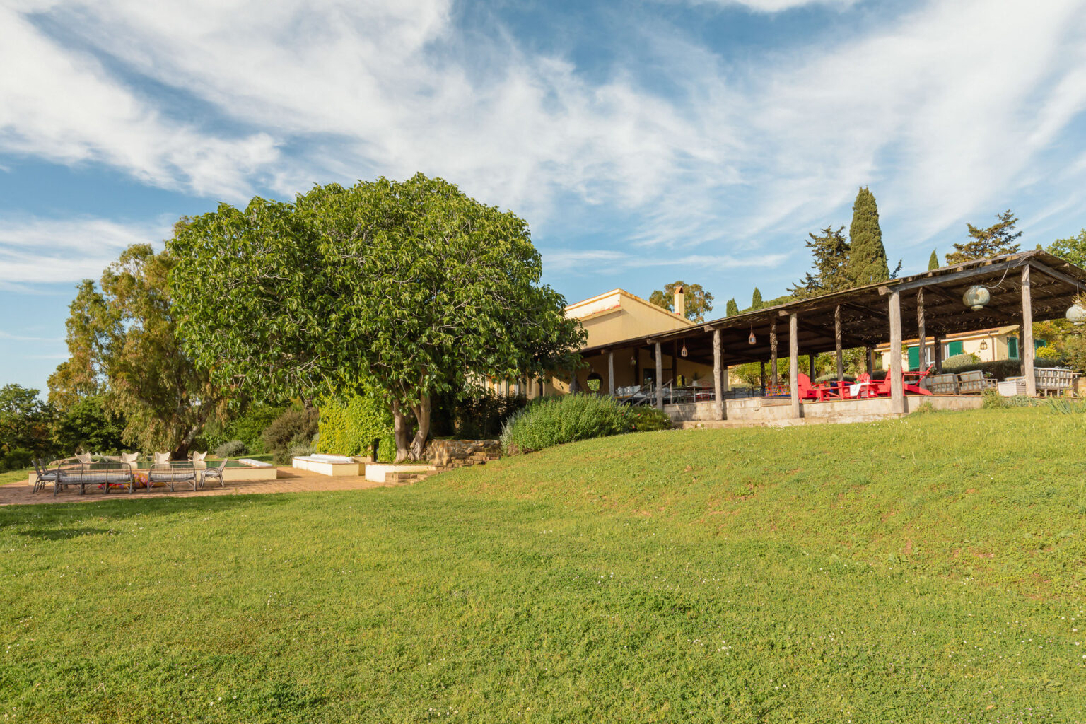 Scenic view of a spacious villa with a wooden terrace surrounded by lush greenery and a large tree, set against a clear blue sky. | Only on SopranoVillas