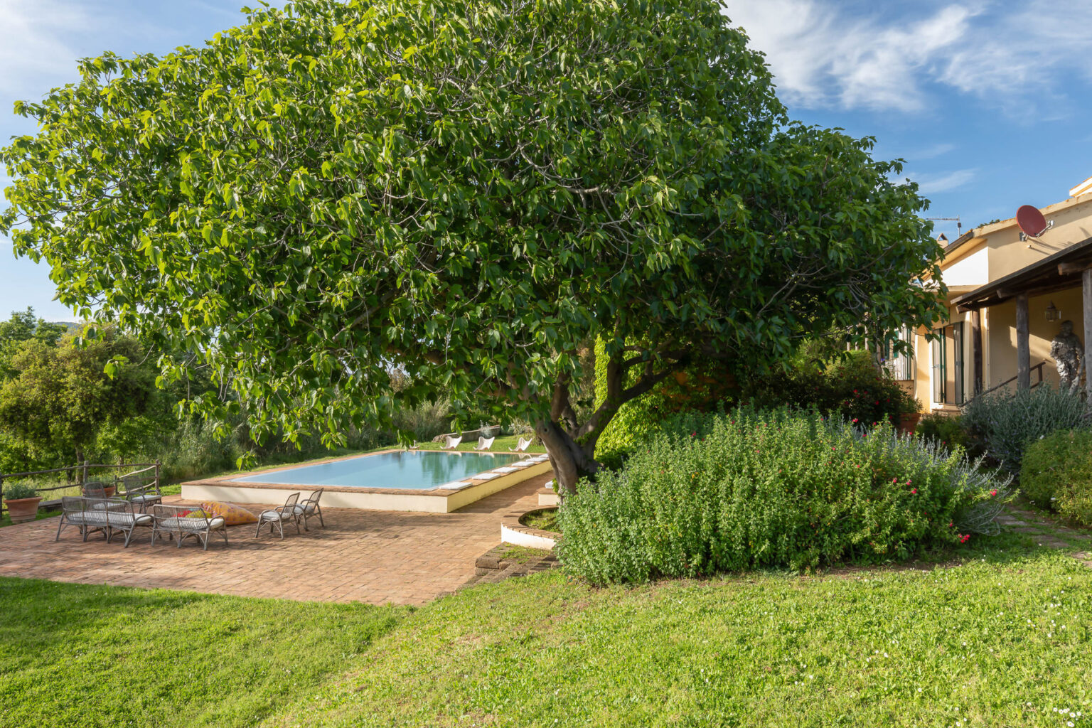 Lush green garden featuring a large tree, a serene swimming pool, and a cozy home under a clear blue sky. Ideal for relaxation and outdoor living. | Only on SopranoVillas