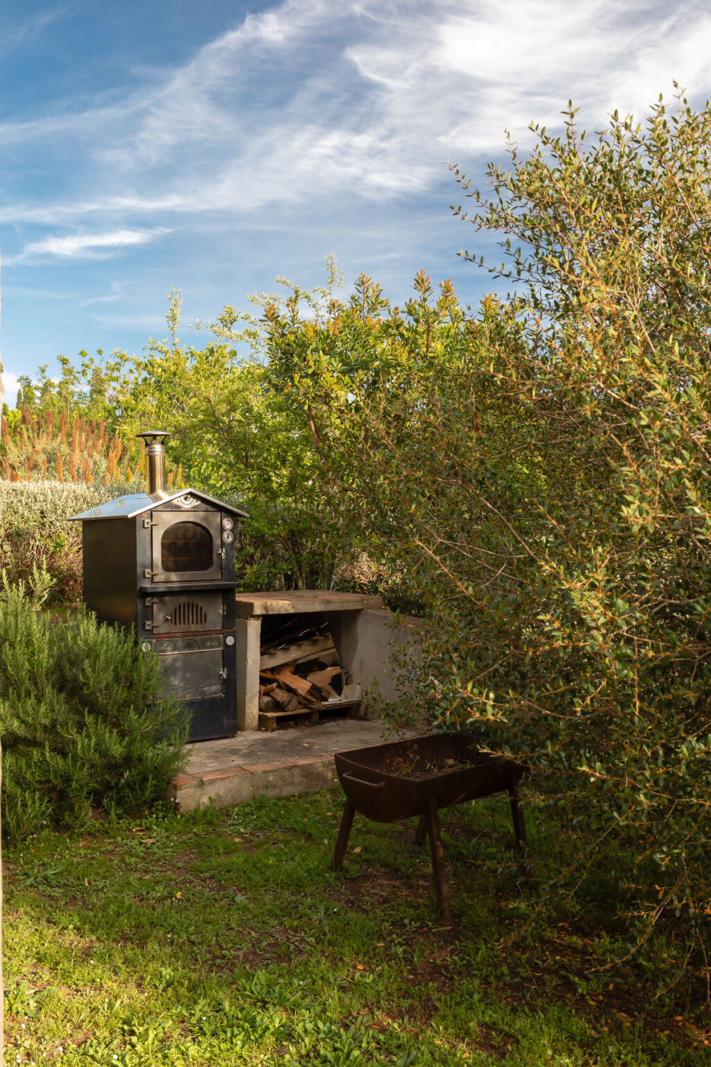 Outdoor pizza oven surrounded by greenery, with a wood storage area nearby, ideal for backyard cooking and entertaining. | Only on SopranoVillas