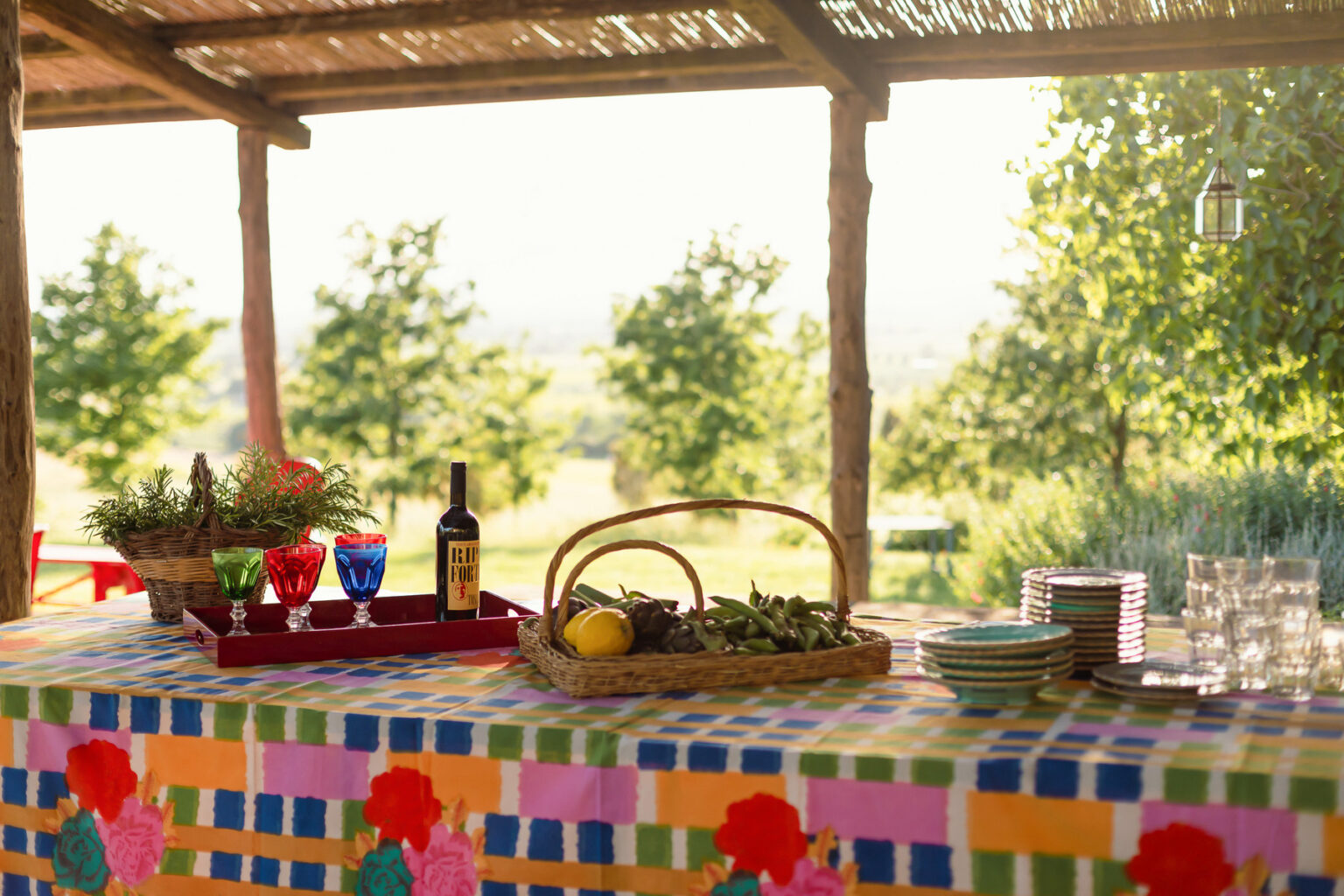 Colorful outdoor dining setup featuring a checkered tablecloth, a basket of fresh greens, a bottle of wine, and vibrant glasses, with a scenic view of greenery in the background. | Only on SopranoVillas