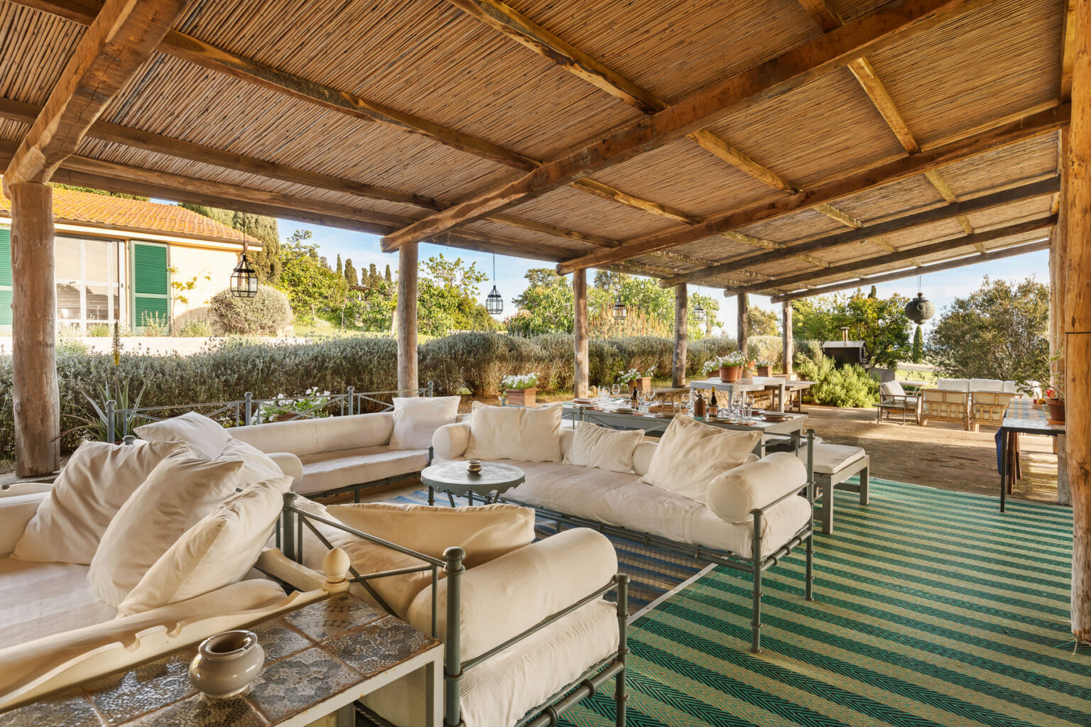 Spacious outdoor living area featuring plush white sofas, a decorative coffee table, and a rustic wooden ceiling. Lush greenery and potted plants enhance the serene atmosphere, with a dining setup visible in the background, perfect for entertaining guests. | Only on SopranoVillas