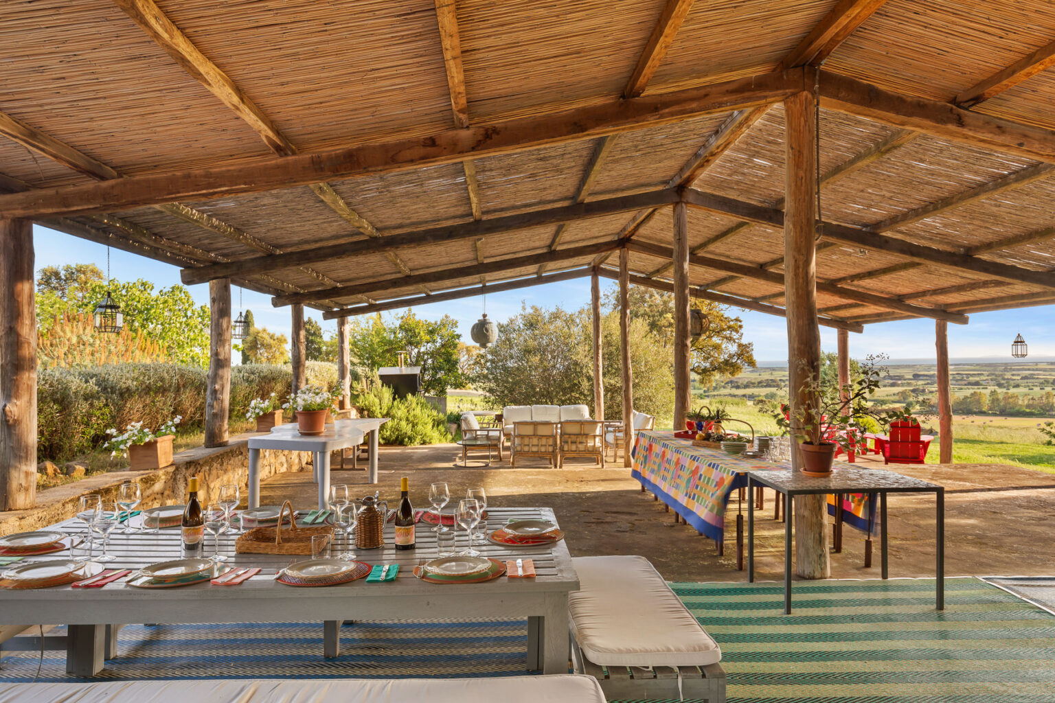 Outdoor dining area with a rustic wooden roof, featuring a long table set with plates, wine bottles, and glasses, surrounded by lush greenery and scenic views. Cozy seating arrangements and colorful table decor enhance the inviting atmosphere, perfect for gatherings and meals in nature. | Only on SopranoVillas