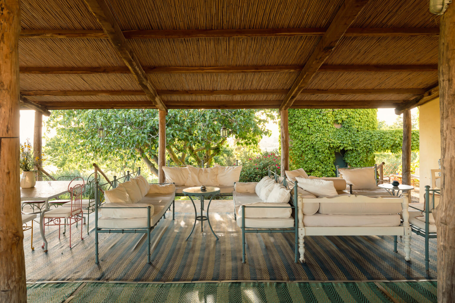Cozy outdoor seating area with comfortable couches and a glass table, surrounded by lush greenery and wooden beams, ideal for relaxation and entertaining. | Only on SopranoVillas