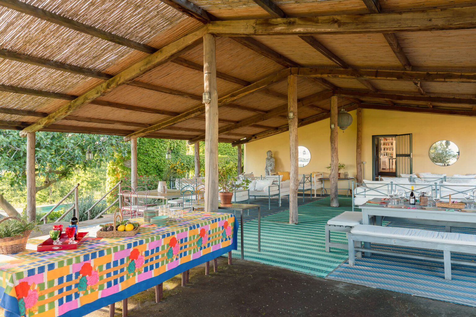 Outdoor dining area with a colorful tablecloth, surrounded by lush greenery, featuring wooden seating and a rustic roof structure, ideal for gatherings and events. | Only on SopranoVillas