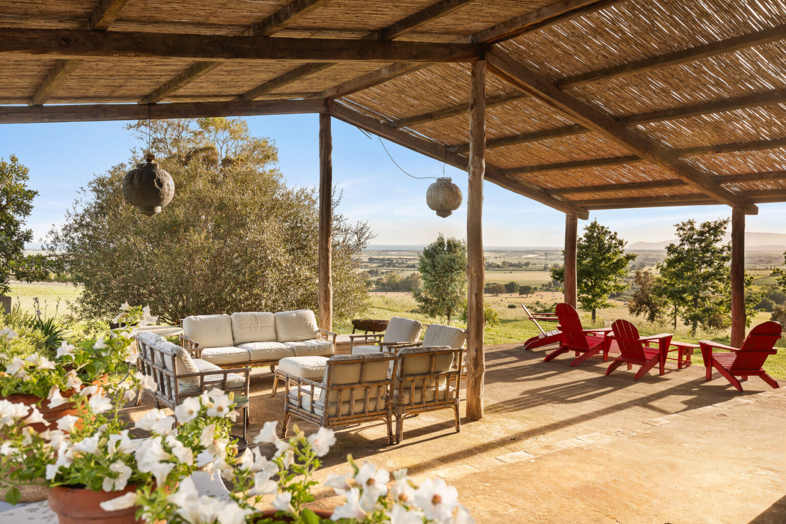 Outdoor patio with comfortable seating and red lounge chairs, surrounded by lush greenery and scenic views, featuring a thatched roof for shade and blooming flowers in the foreground. | Only on SopranoVillas