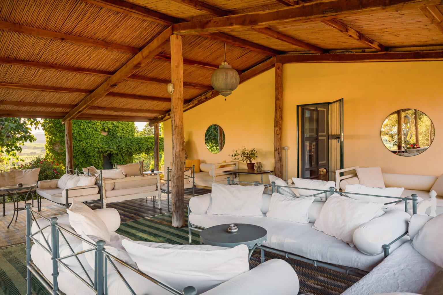 Cozy outdoor lounge area with comfortable white sofas and a round table, set under a wooden roof surrounded by greenery, ideal for relaxation and social gatherings. | Only on SopranoVillas