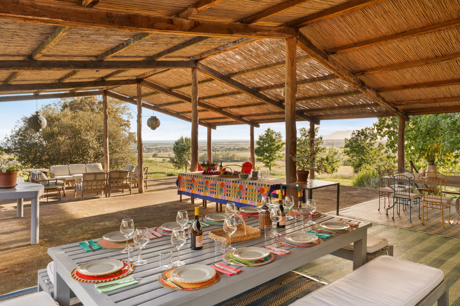 Outdoor dining area with a rustic wooden table set for a meal, featuring colorful plates, glasses, and wine bottles. The space is covered with a thatched roof, surrounded by lush greenery and a scenic landscape in the background, creating a cozy and inviting atmosphere for gatherings. | Only on SopranoVillas