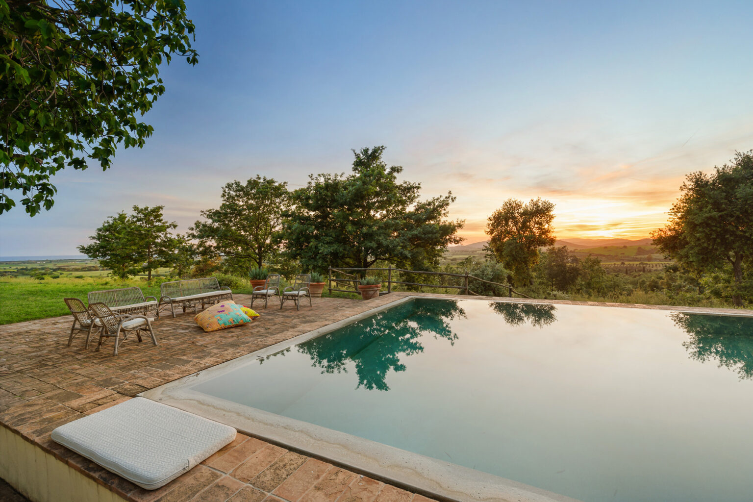 Scenic view of a tranquil infinity pool surrounded by lush greenery at sunset, featuring outdoor seating and vibrant cushions, ideal for relaxation and leisure. | Only on SopranoVillas