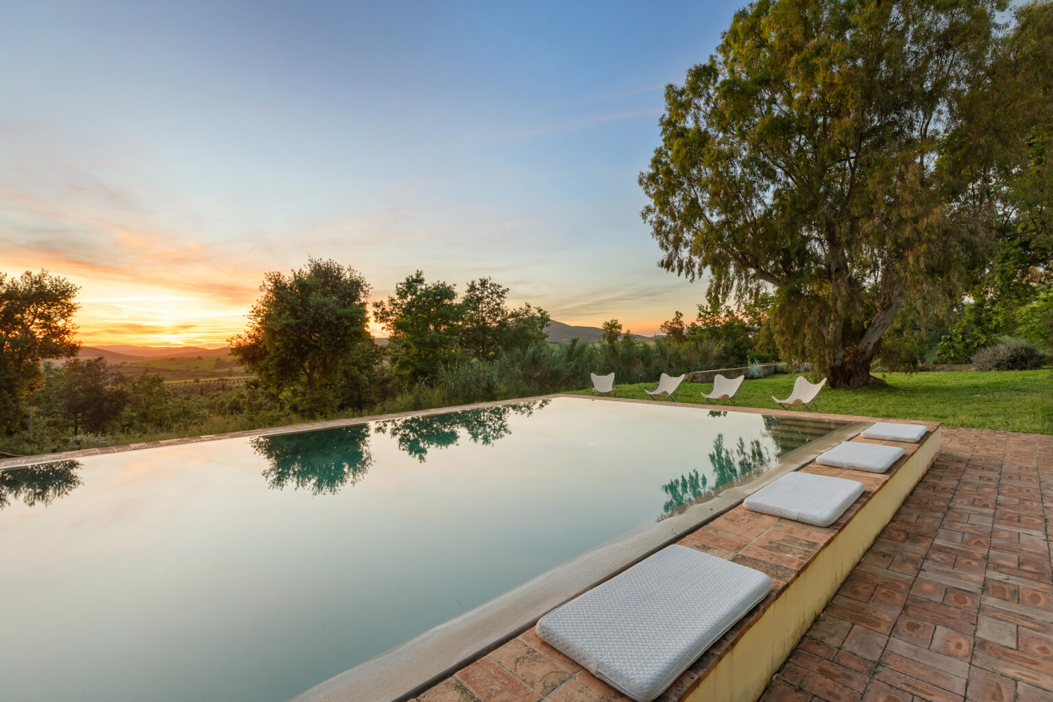 Scenic view of a serene swimming pool surrounded by lush greenery at sunset, featuring comfortable lounge chairs along the edge. | Only on SopranoVillas