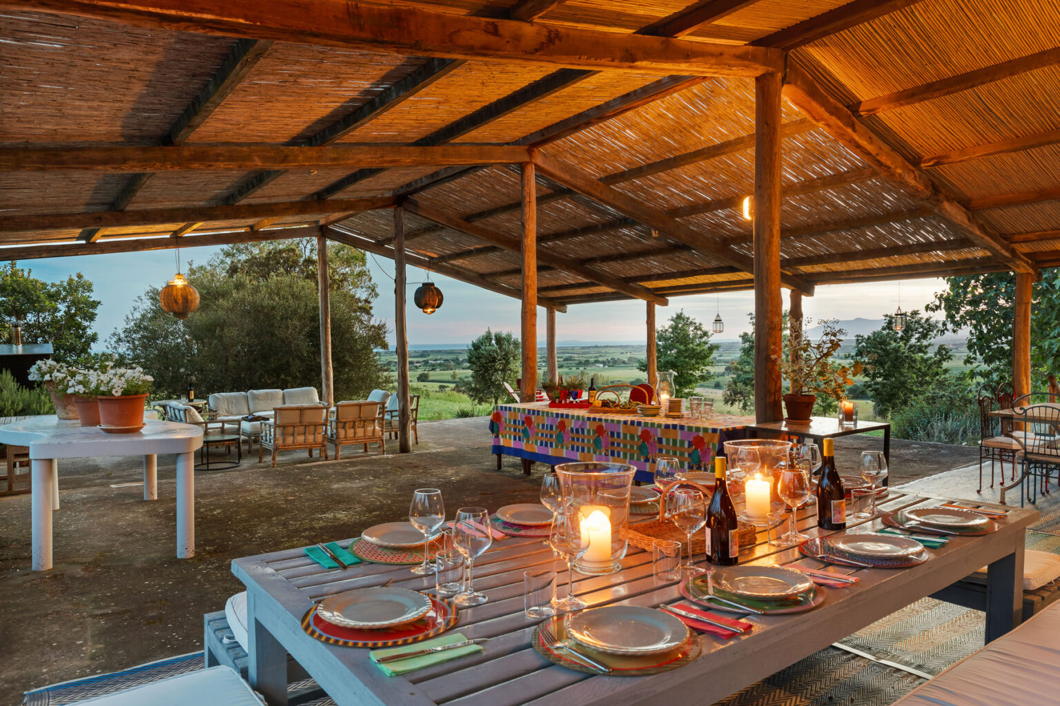Outdoor dining area with rustic wooden tables set for a meal, featuring colorful tableware and candles, under a thatched roof with scenic views of nature at sunset. | Only on SopranoVillas