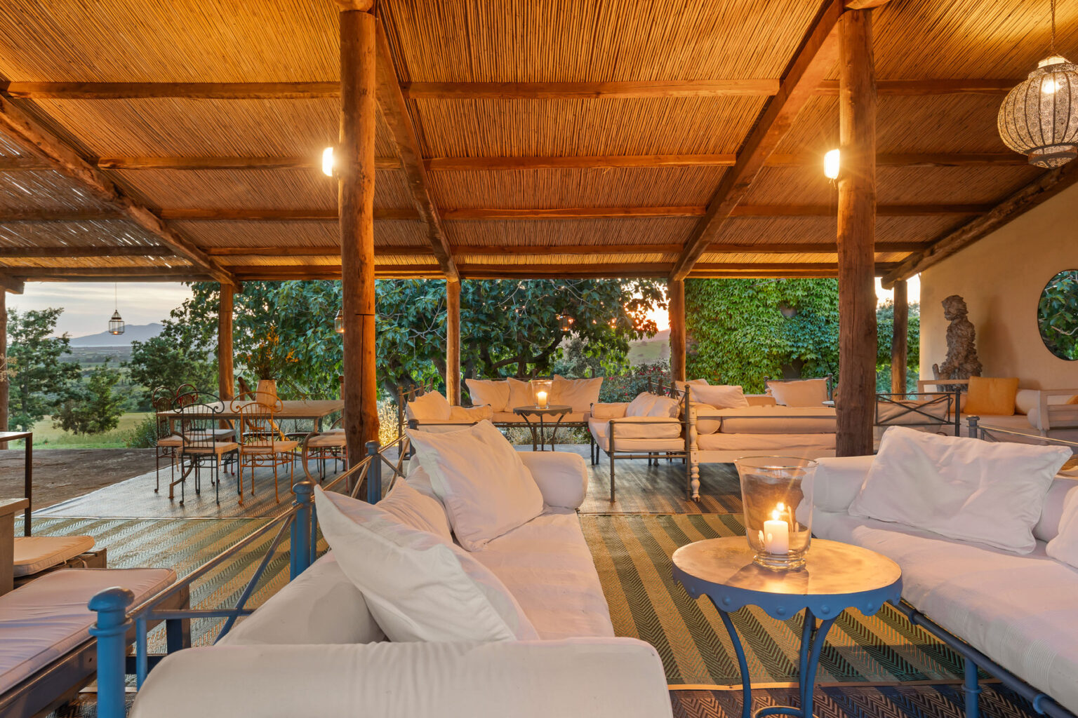 Cozy outdoor lounge area with comfortable white seating, wooden beams, and warm lighting, surrounded by greenery and offering a serene atmosphere for relaxation. | Only on SopranoVillas