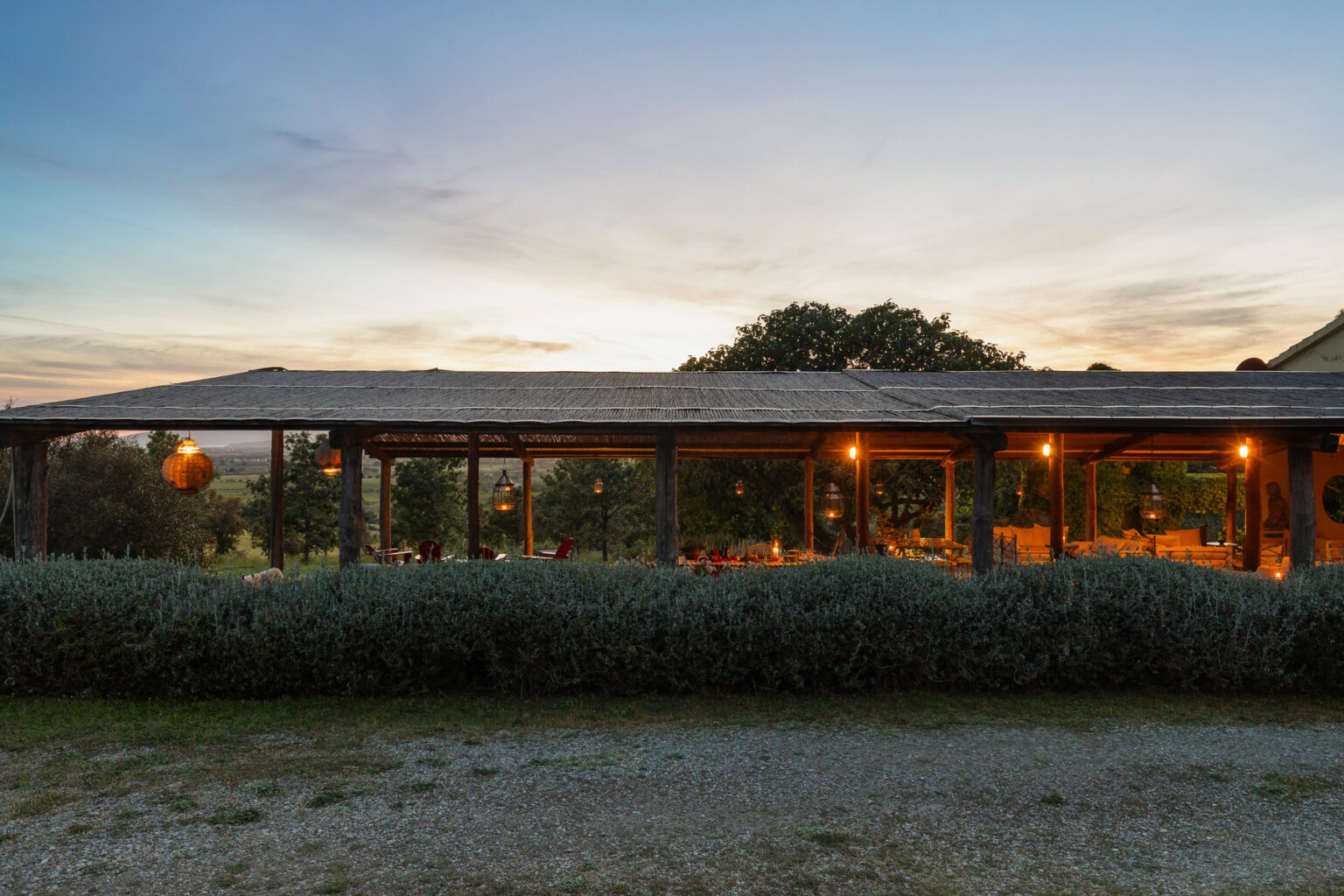 Rustic outdoor dining area with wooden structure, warm lighting, and scenic sunset backdrop, surrounded by greenery and gravel path. Ideal for cozy gatherings. | Only on SopranoVillas