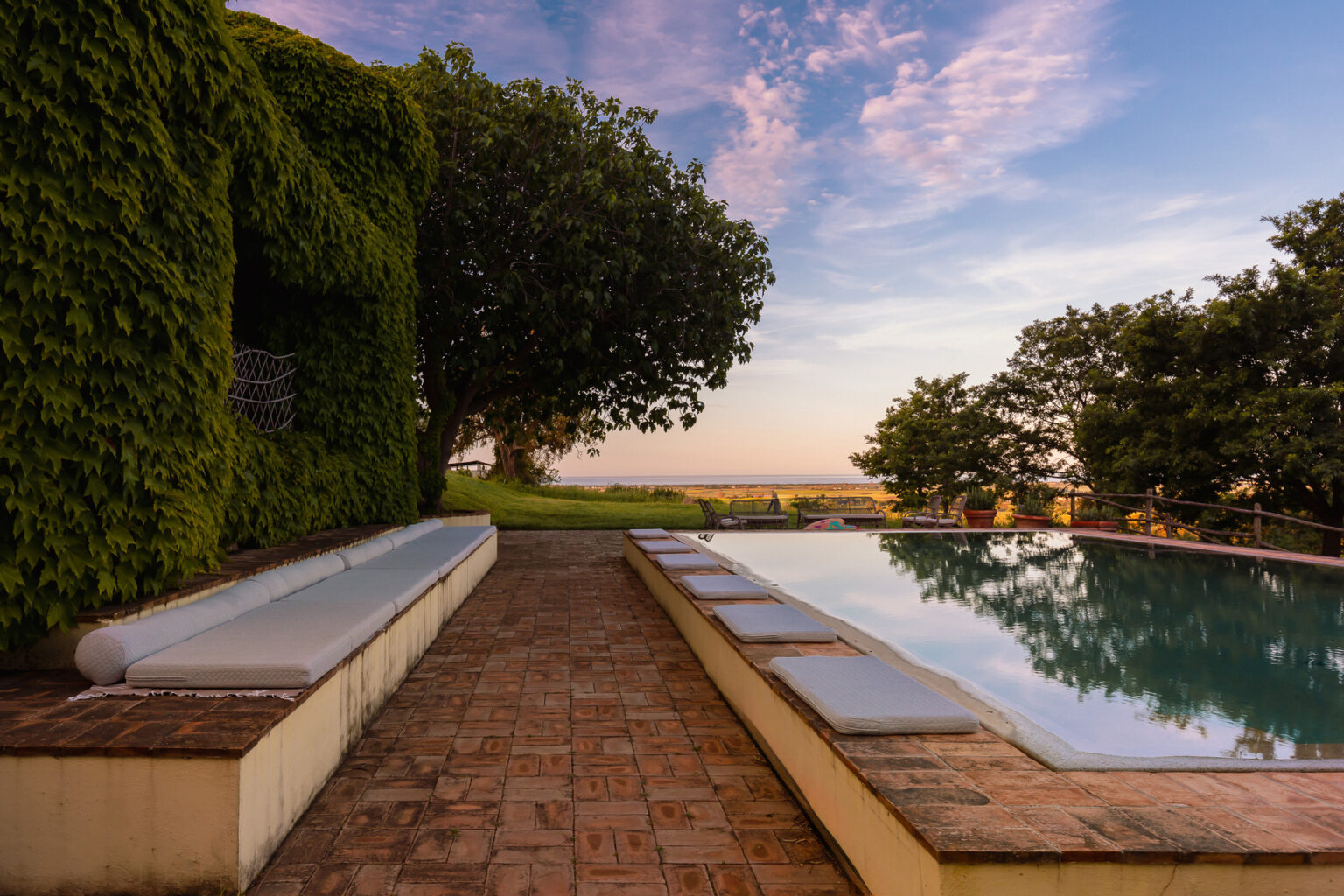 Scenic outdoor pool area surrounded by lush greenery and trees, featuring a spacious patio with lounge seating, under a vibrant sunset sky. | Only on SopranoVillas