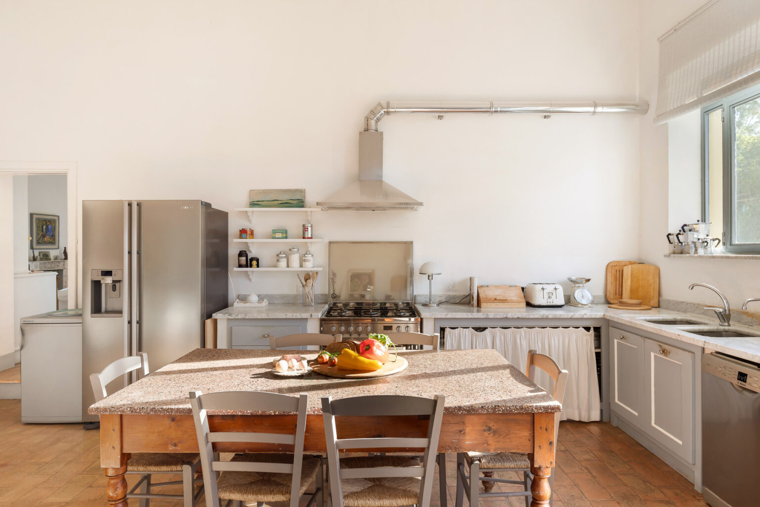 Bright and spacious modern kitchen featuring a wooden dining table with a fruit centerpiece, stainless steel appliances, and sleek cabinetry, illuminated by natural light from large windows. | Only on SopranoVillas