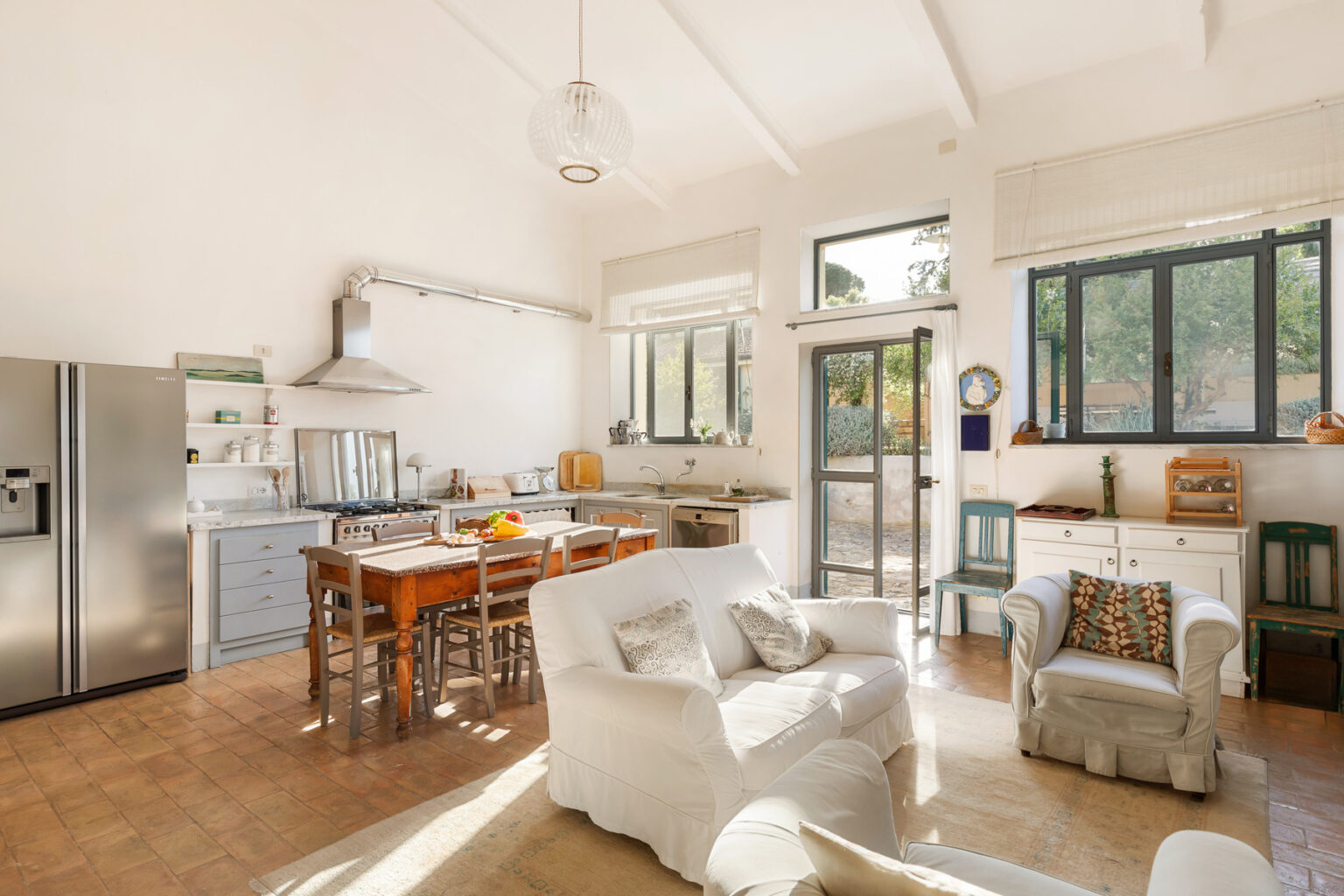 Bright and airy kitchen and living area featuring white furniture, a wooden dining table, stainless steel appliances, and large windows that invite natural light. | Only on SopranoVillas