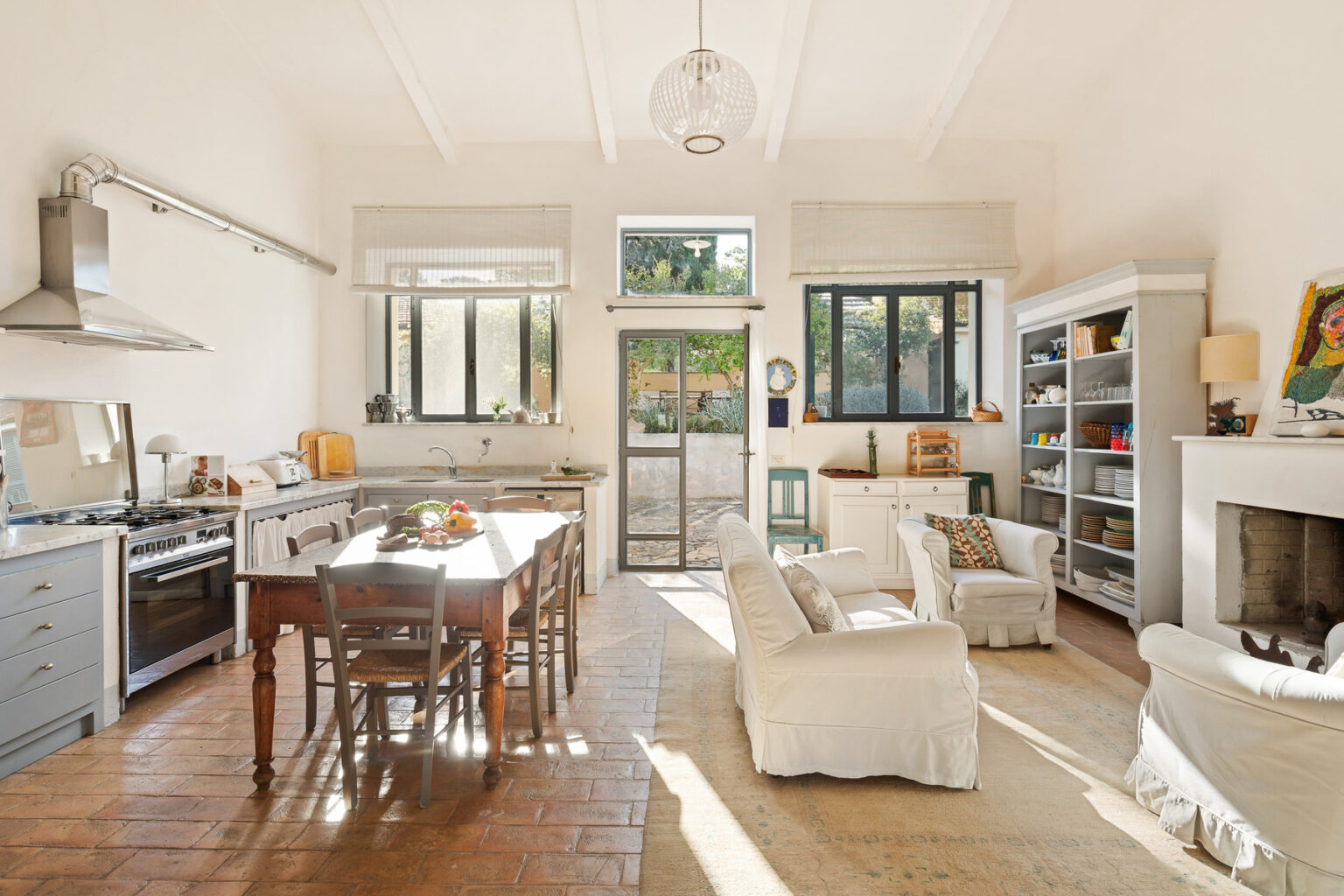 Bright and airy modern kitchen and dining area featuring a large wooden table, white chairs, and a cozy seating area. Sunlight streams through large windows, highlighting the stylish appliances and open layout. | Only on SopranoVillas