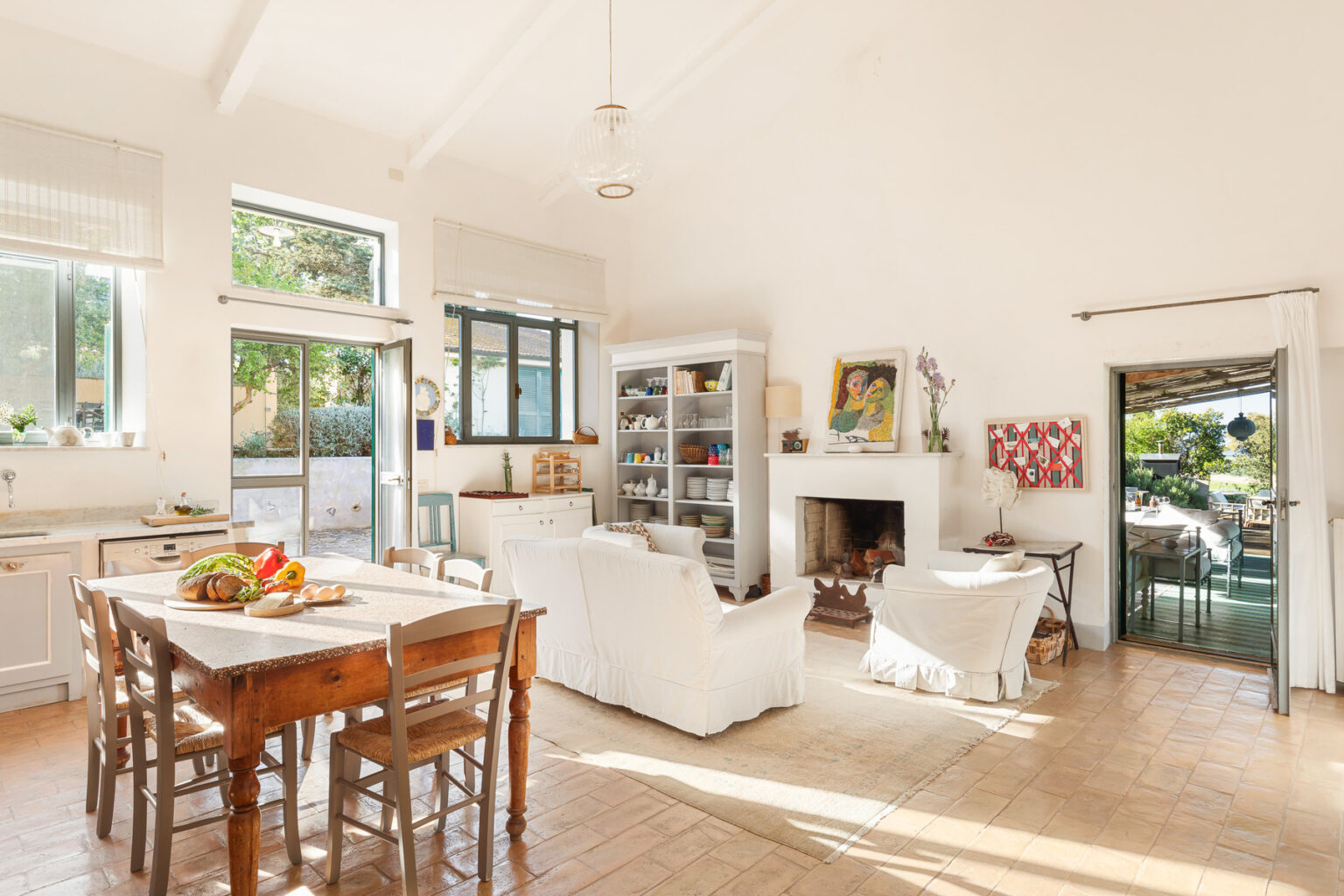 Bright and airy living room featuring a white color scheme, large windows, and a cozy fireplace. The space includes a wooden dining table with chairs, a comfortable sofa, and decorative artwork on the walls, creating a welcoming atmosphere. | Only on SopranoVillas