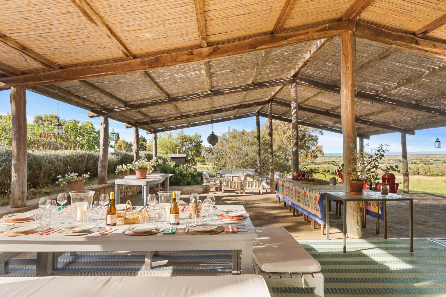 Outdoor dining area with a rustic wooden structure, featuring a long table set for a meal, surrounded by greenery and scenic views. The space is decorated with potted plants, glassware, and colorful table settings, creating an inviting atmosphere for gatherings. | Only on SopranoVillas