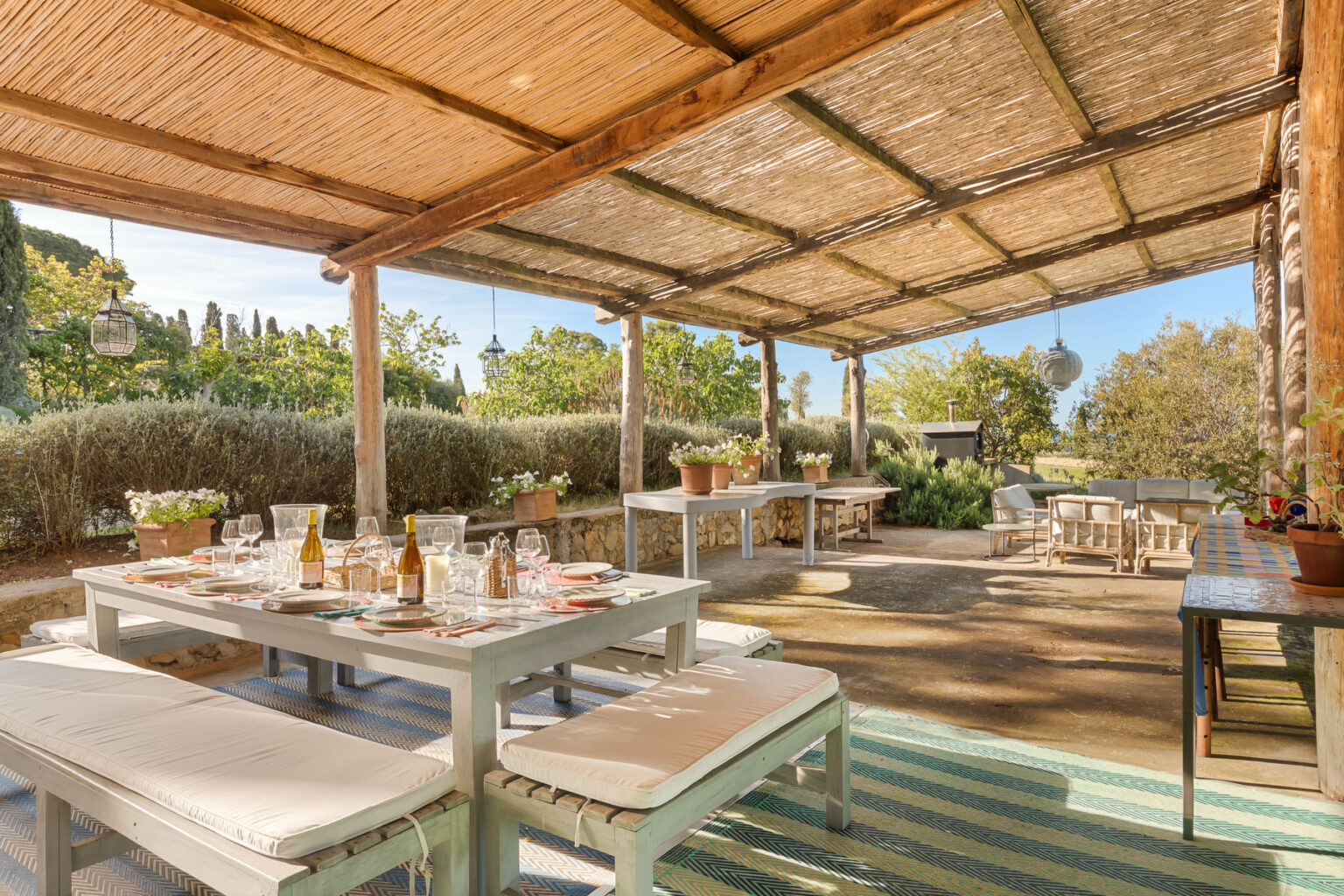 Outdoor dining area featuring a rustic wooden structure with a thatched roof, set up with a large table adorned with wine bottles, glasses, and decorative plates. Surrounded by greenery and potted plants, the space offers a relaxed atmosphere for gatherings, complemented by comfortable seating and ambient lighting. | Only on SopranoVillas