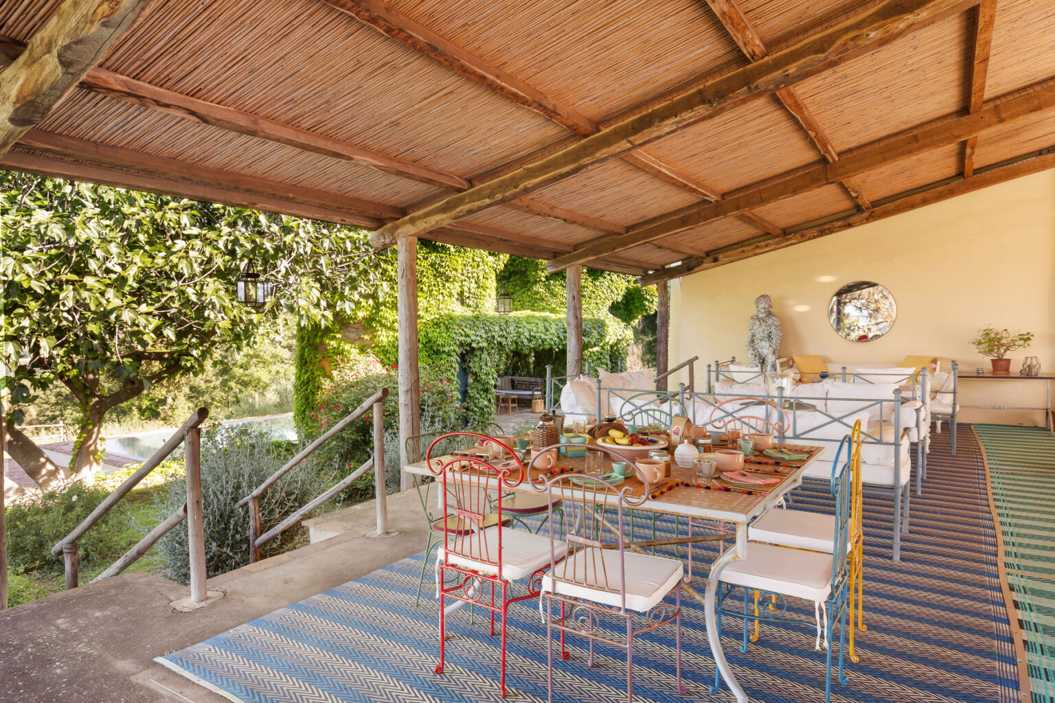 Outdoor dining area featuring a rustic table set with dishes and fruit, surrounded by colorful metal chairs. The space is shaded by a thatched roof and adorned with greenery, creating a cozy atmosphere. A decorative statue and a mirror enhance the aesthetic, while a patterned rug adds warmth to the setting. | Only on SopranoVillas