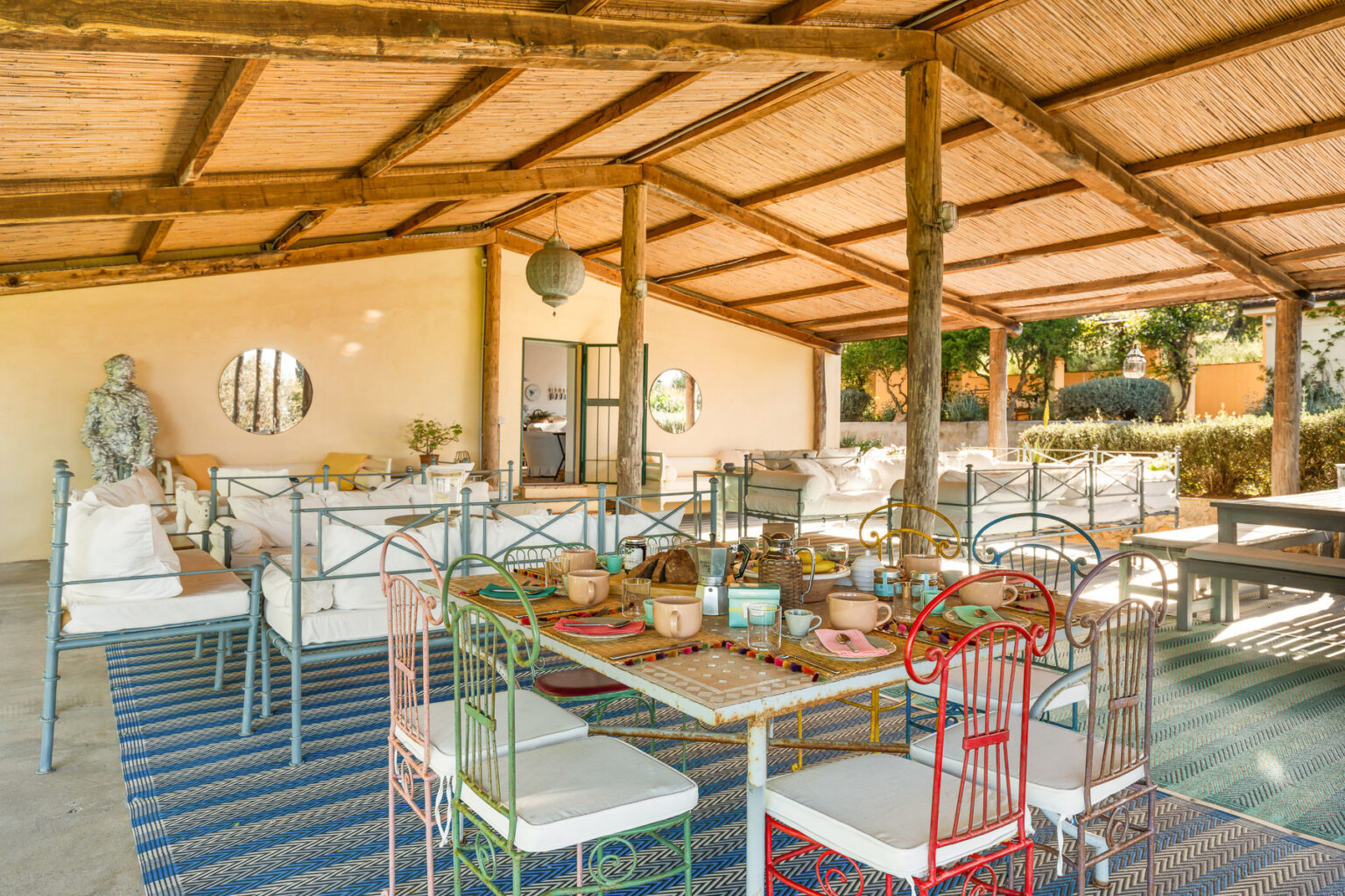 Stylish outdoor dining area featuring colorful metal chairs, a rustic wooden ceiling, and a cozy seating arrangement with plush cushions. The table is beautifully set with various dishes and drinks, surrounded by lush greenery, creating an inviting atmosphere for gatherings. | Only on SopranoVillas
