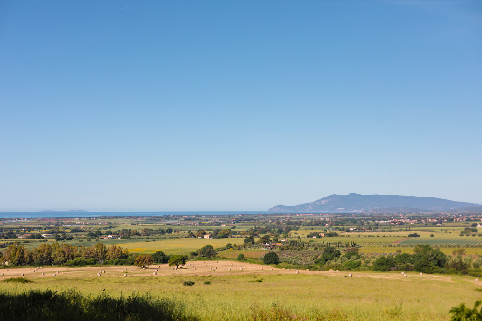Panoramic view of lush green fields and distant mountains under a clear blue sky, with the ocean visible on the horizon, showcasing the natural beauty of the landscape. | Only on SopranoVillas