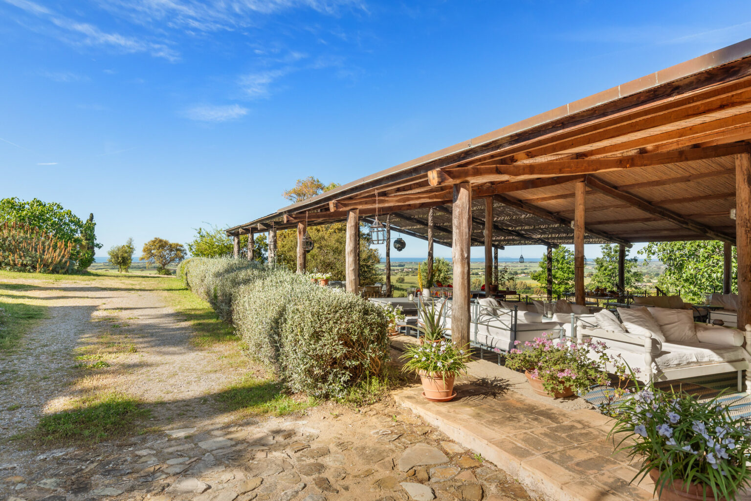 Rustic outdoor seating area with wooden beams, surrounded by greenery and a gravel path, offering a serene view of the landscape under a clear blue sky. | Only on SopranoVillas
