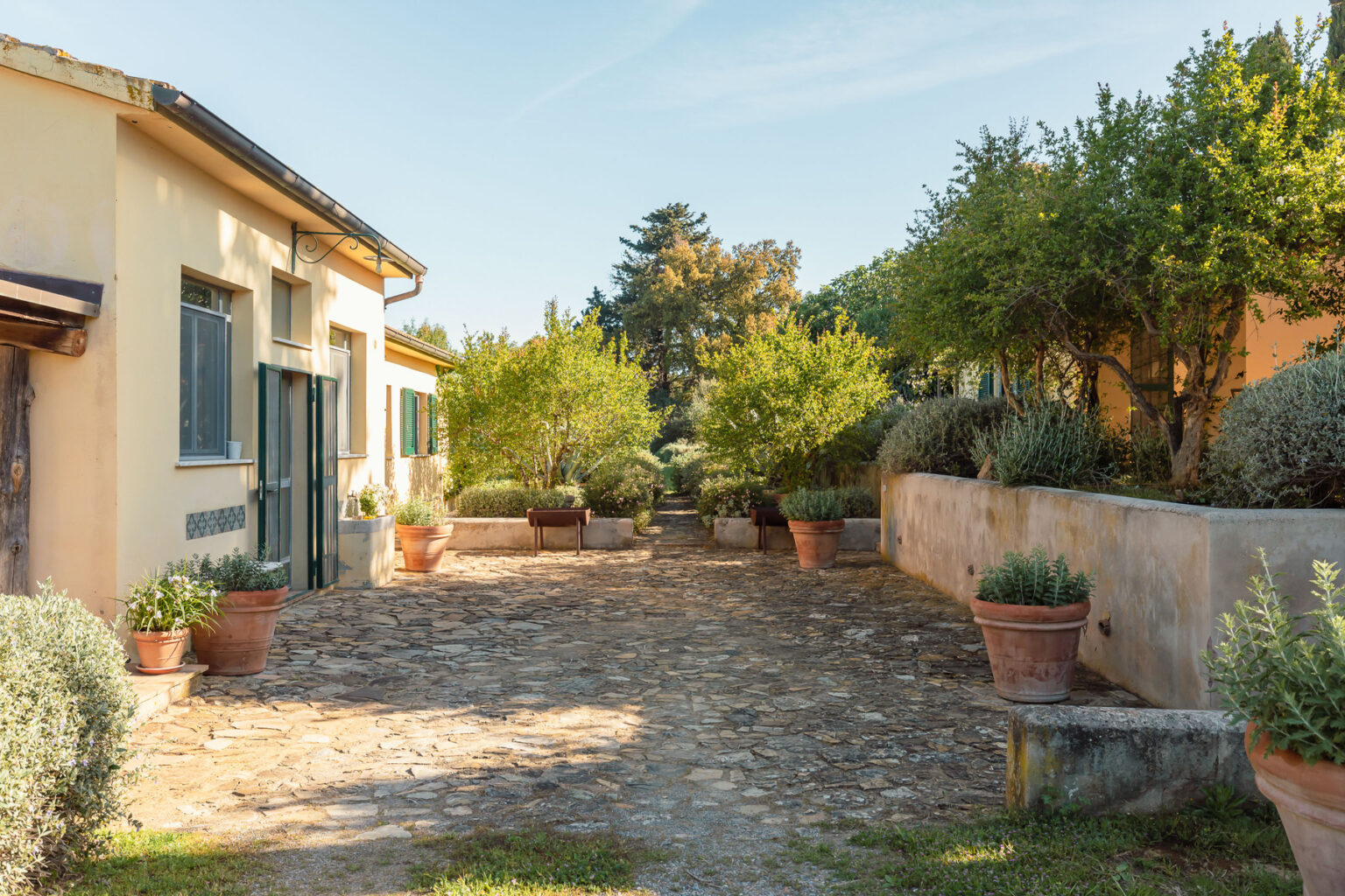 Charming courtyard with stone pathway, surrounded by lush greenery and potted plants, featuring a rustic building with large windows under a clear blue sky. | Only on SopranoVillas