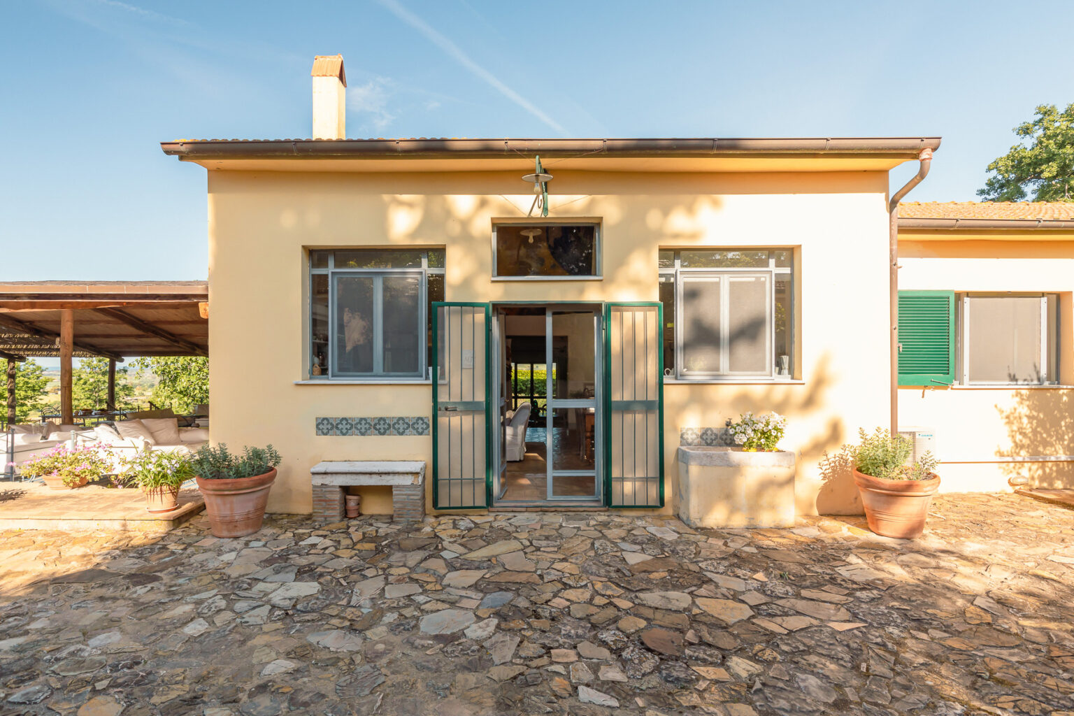 Charming yellow house with green shutters and an inviting entrance, surrounded by a stone pathway and potted plants, set against a clear blue sky. Ideal for showcasing rustic architecture and outdoor living spaces. | Only on SopranoVillas