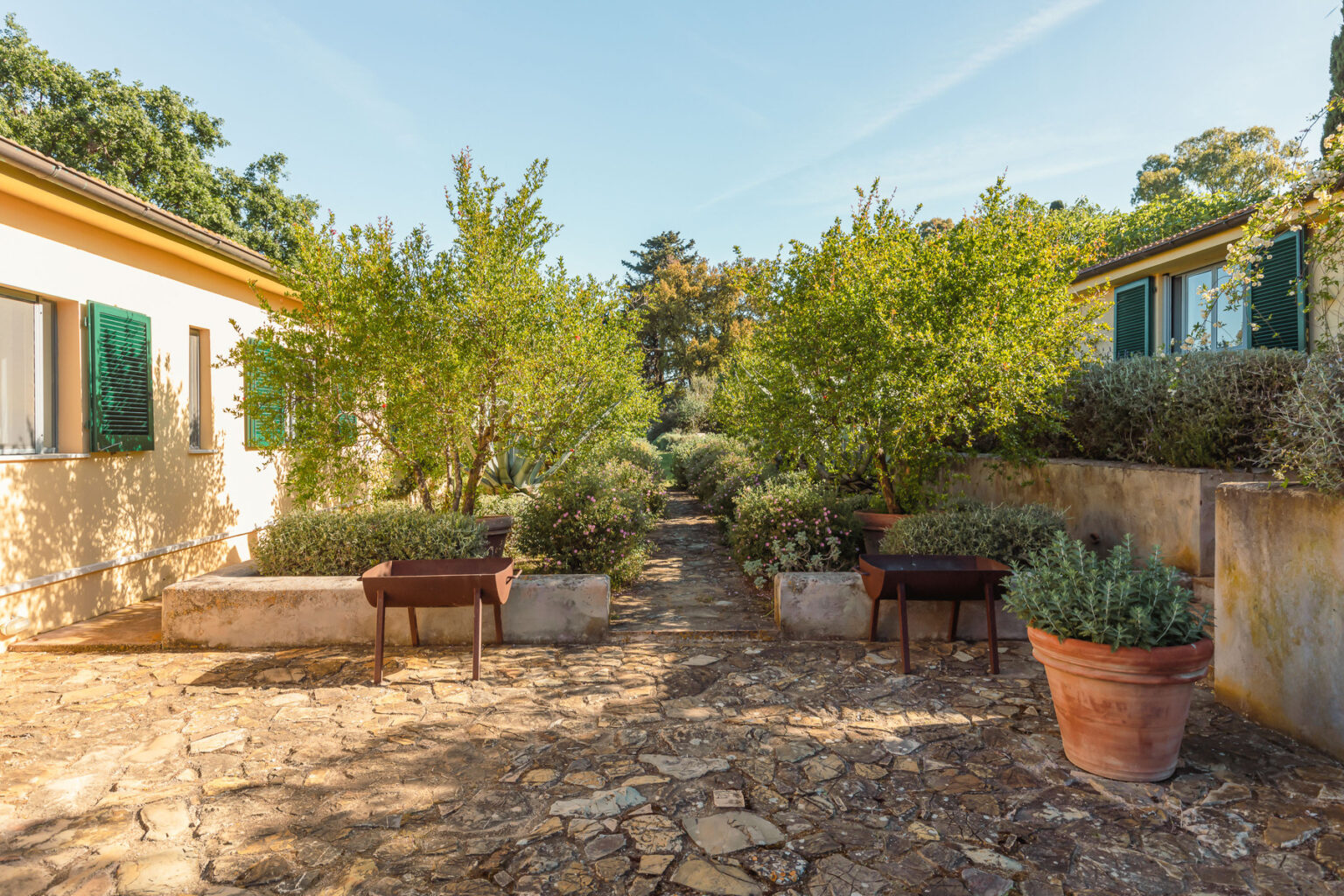 Lush garden pathway flanked by potted plants and trees, leading to cozy cottages with green shutters, under a clear blue sky. | Only on SopranoVillas