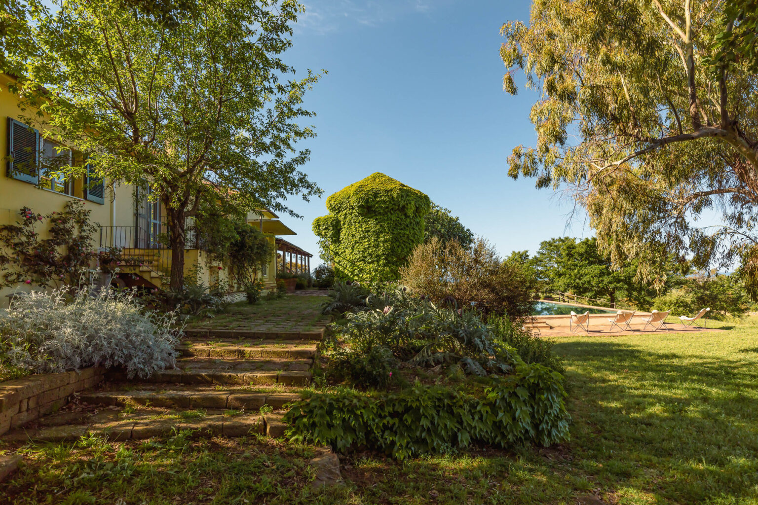 Lush garden landscape with greenery and well-maintained pathways leading to a charming yellow house under a clear blue sky. | Only on SopranoVillas
