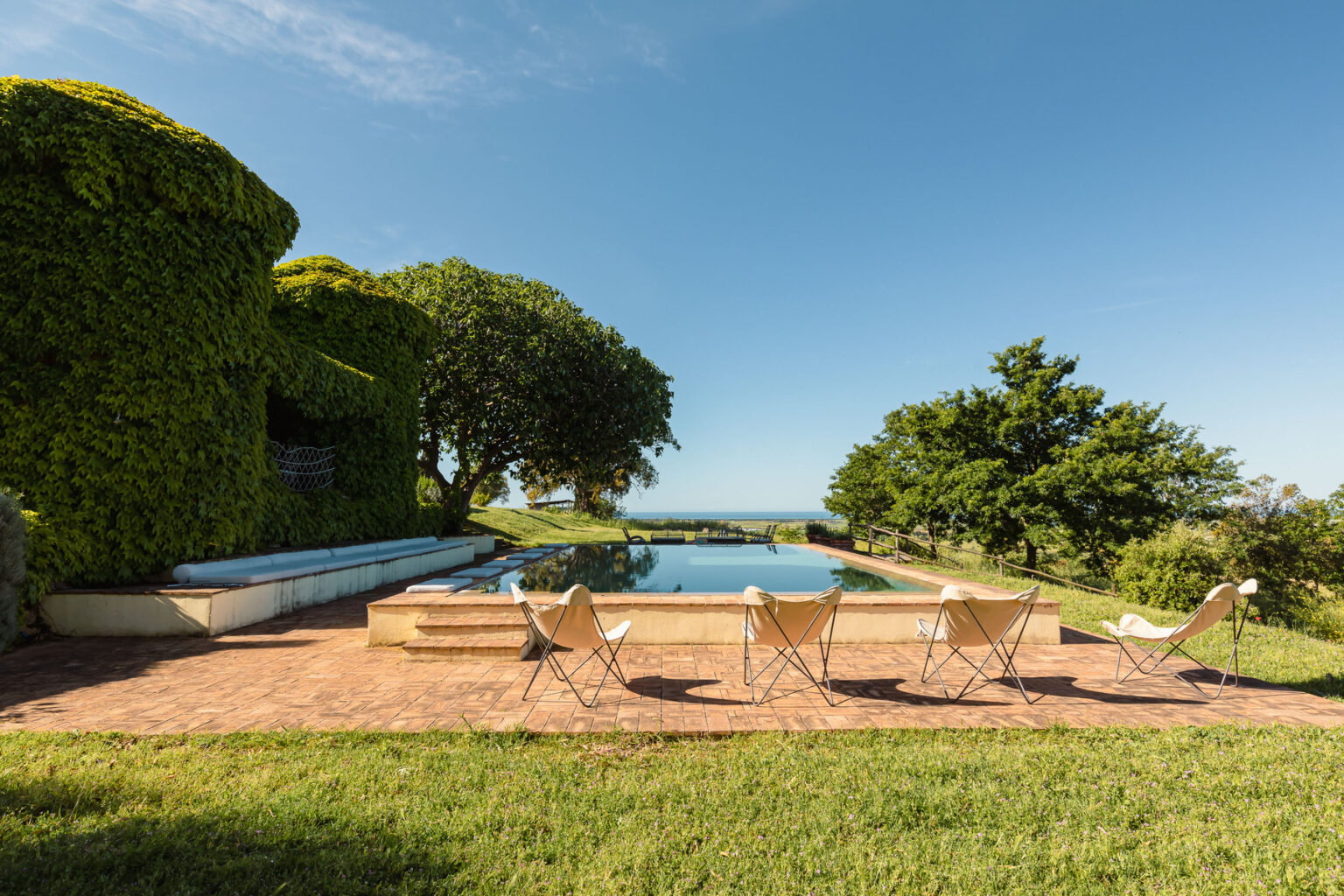 Modern outdoor pool area with lounge chairs, surrounded by lush greenery and trees, overlooking a scenic view under a clear blue sky. | Only on SopranoVillas
