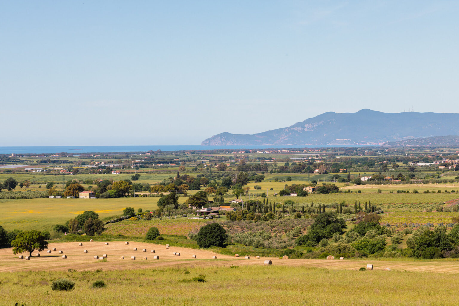 Scenic landscape view of rolling green hills, fields, and distant mountains overlooking a calm sea, showcasing the beauty of nature and agricultural land in a coastal region. | Only on SopranoVillas