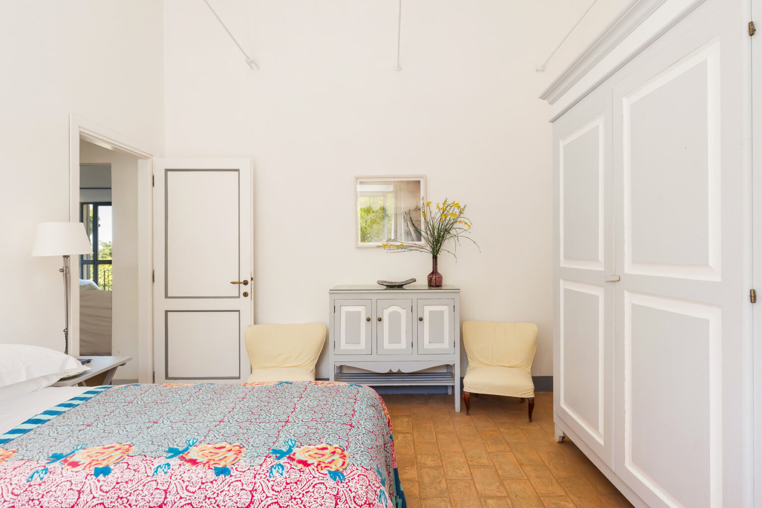 Bright and airy bedroom featuring a colorful floral bedspread, two cream chairs, a wooden cabinet with a vase of flowers, and large windows allowing natural light. The minimalist design includes white walls and wooden flooring, creating a serene atmosphere. | Only on SopranoVillas