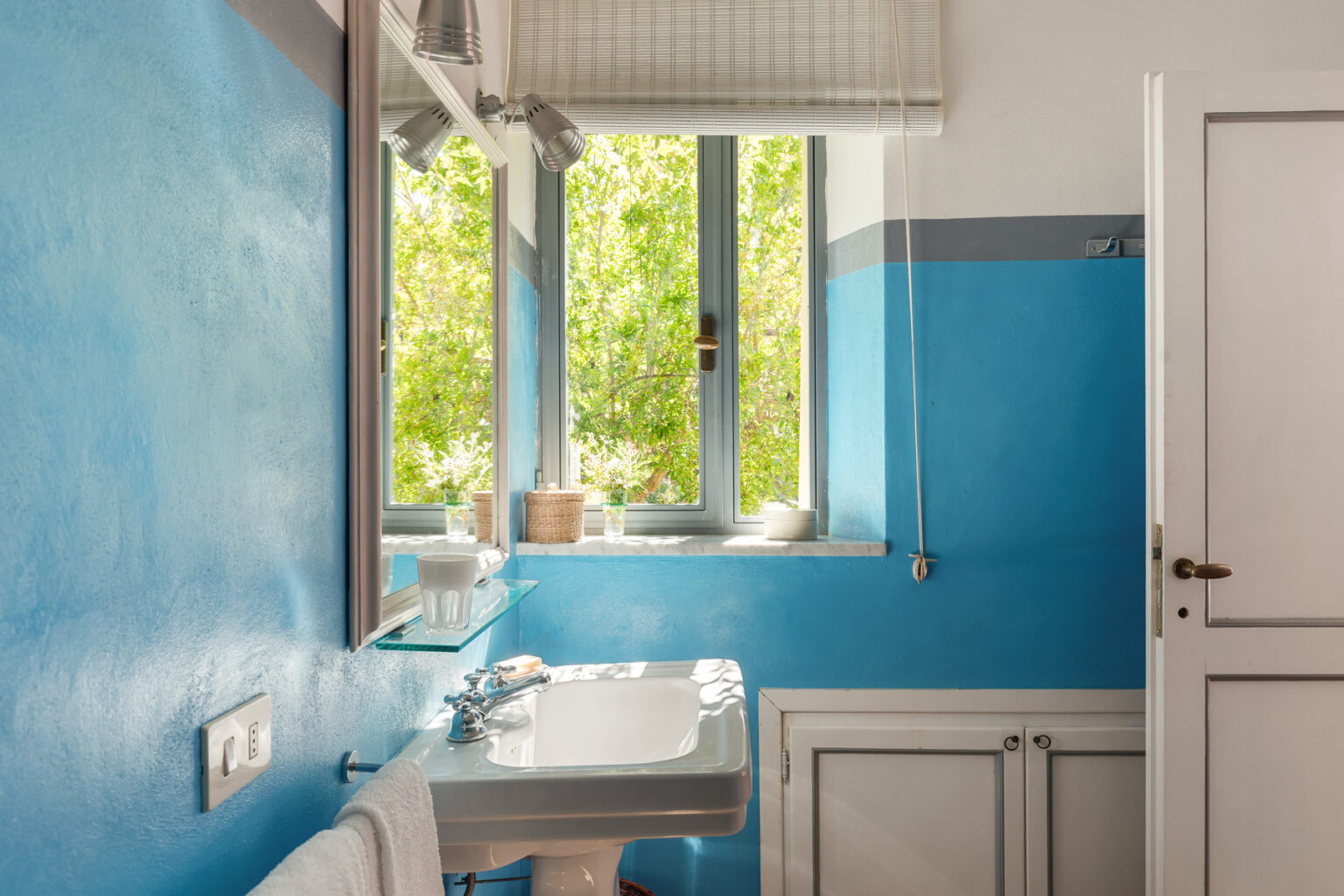 Bright and modern bathroom featuring blue walls, a white sink, and large windows allowing natural light, complemented by greenery outside. The design includes a mirror, glass shelves, and stylish decor elements, creating a fresh and inviting atmosphere. | Only on SopranoVillas