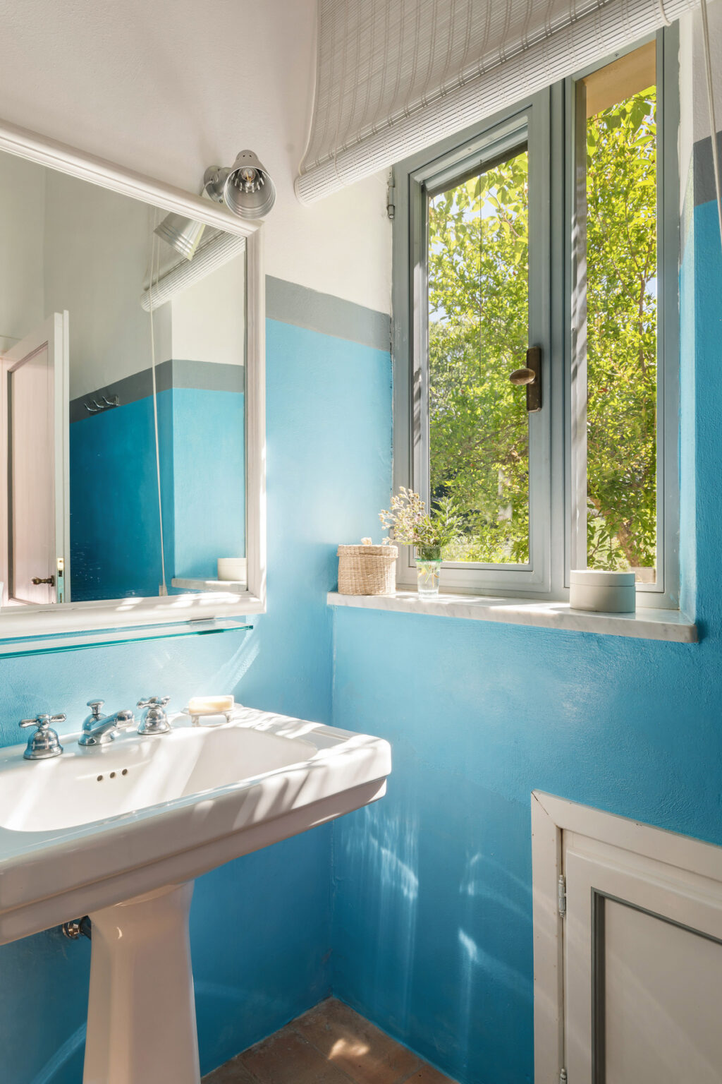Bright and airy bathroom featuring a white sink, a large mirror, and blue walls with a gray stripe. Sunlight streams through a window adorned with a light shade, highlighting a small decorative plant and a woven basket on the windowsill. The space exudes a fresh and inviting atmosphere. | Only on SopranoVillas