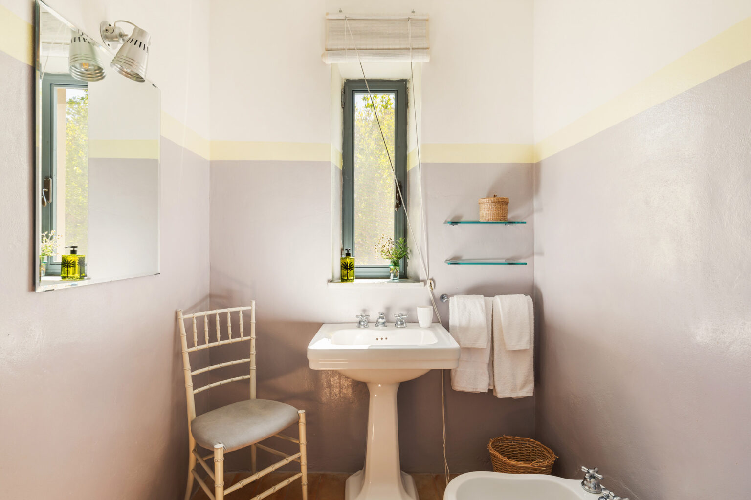 Cozy bathroom featuring a vintage pedestal sink, a large mirror, and a comfortable chair. Soft gray walls with a yellow accent stripe create a calming atmosphere. Natural light streams in through a window, highlighting decorative items like towels and plants on glass shelves. | Only on SopranoVillas