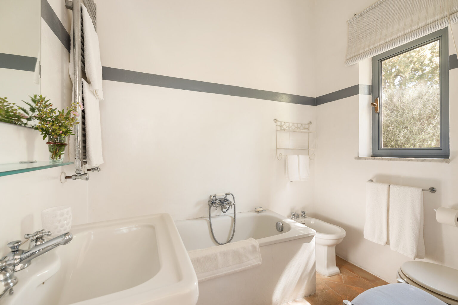 Bright and airy bathroom featuring a white bathtub, modern fixtures, and a window allowing natural light. Decorated with a small plant and neatly arranged towels, the space exudes a clean and relaxing atmosphere. | Only on SopranoVillas