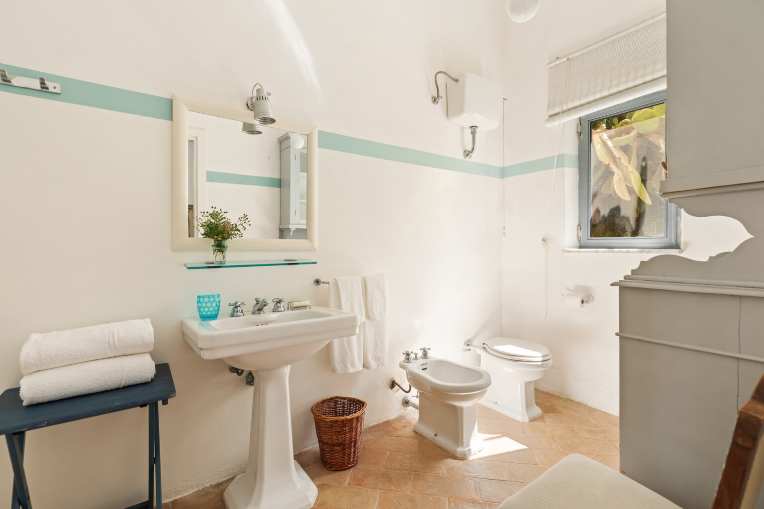 Bright and modern bathroom featuring a white sink, toilet, and a large mirror. The space is accented with light blue stripes and includes neatly stacked towels on a small table. Natural light enters through a window, enhancing the clean and fresh atmosphere. | Only on SopranoVillas