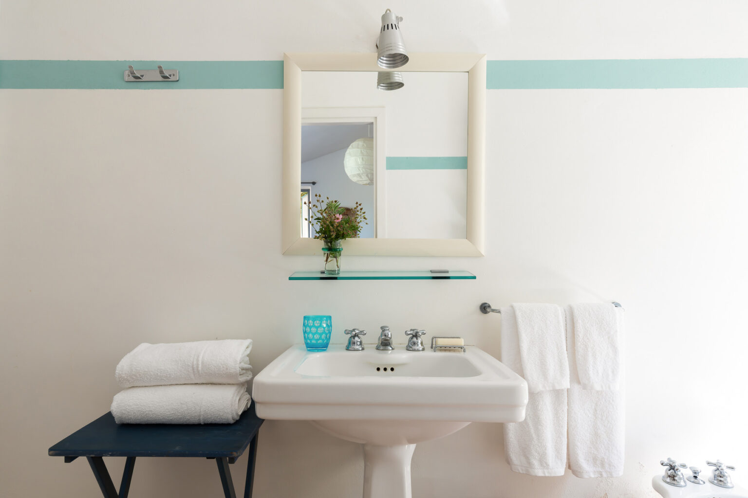 Modern bathroom interior featuring a white sink, a large mirror, neatly stacked white towels, and a small vase with flowers. The walls are accented with light blue stripes, creating a fresh and inviting atmosphere. | Only on SopranoVillas