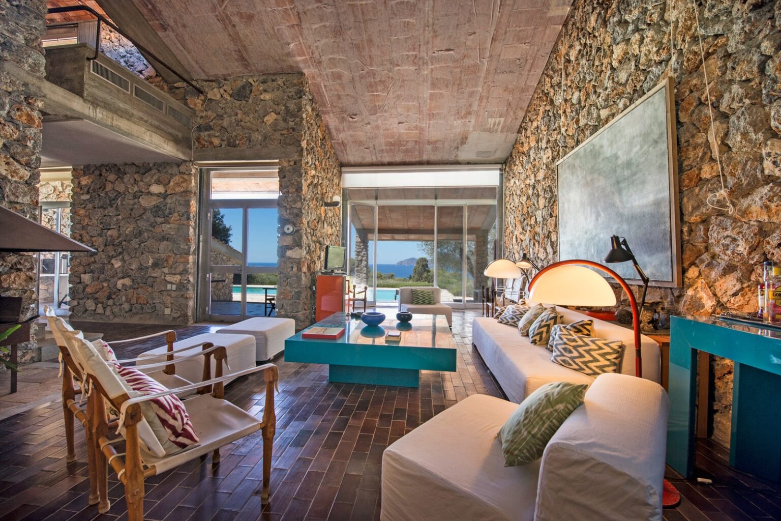 Modern living room with stone walls, large windows, and stylish furnishings. Features a comfortable white sofa with patterned cushions, a turquoise coffee table, and wooden chairs. Natural light floods the space, highlighting the seamless indoor-outdoor connection and views of the landscape. Ideal for contemporary home design inspiration. | Only on SopranoVillas