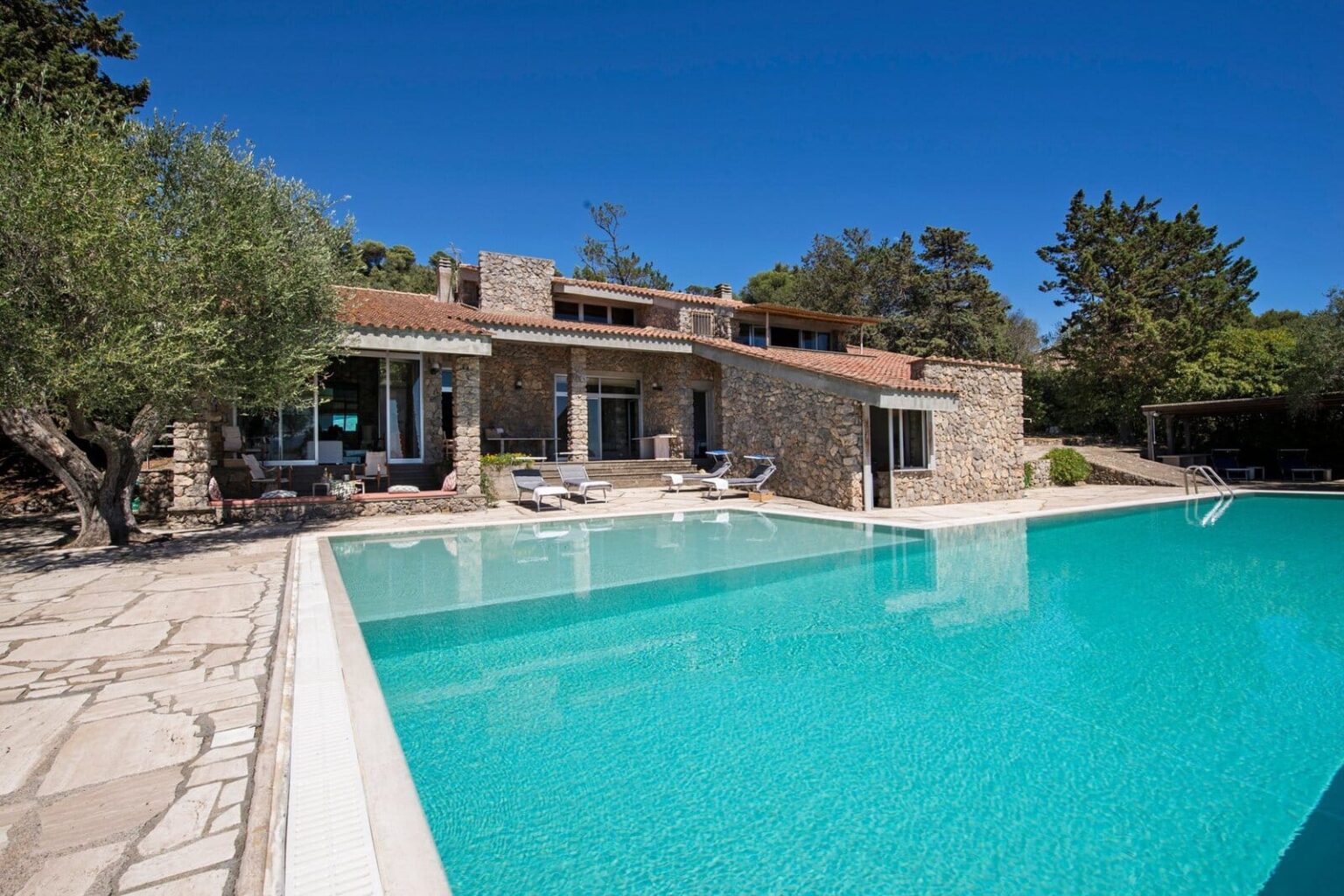 Luxury stone villa with a large swimming pool and sun loungers, surrounded by lush greenery under a clear blue sky. | Only on SopranoVillas