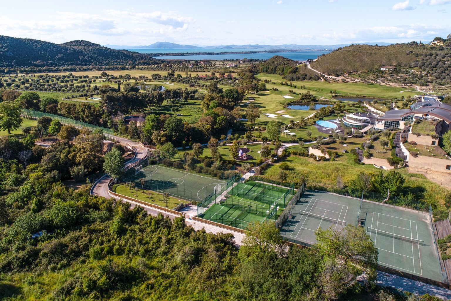 Aerial view of a scenic landscape featuring multiple tennis courts surrounded by lush greenery, rolling hills, and a tranquil body of water in the distance. Ideal setting for outdoor sports and recreation. | Only on SopranoVillas
