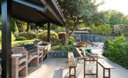 Outdoor kitchen and dining area featuring brick barbecue grills, wooden tables, and lush greenery, ideal for entertaining and alfresco dining in a serene garden setting.