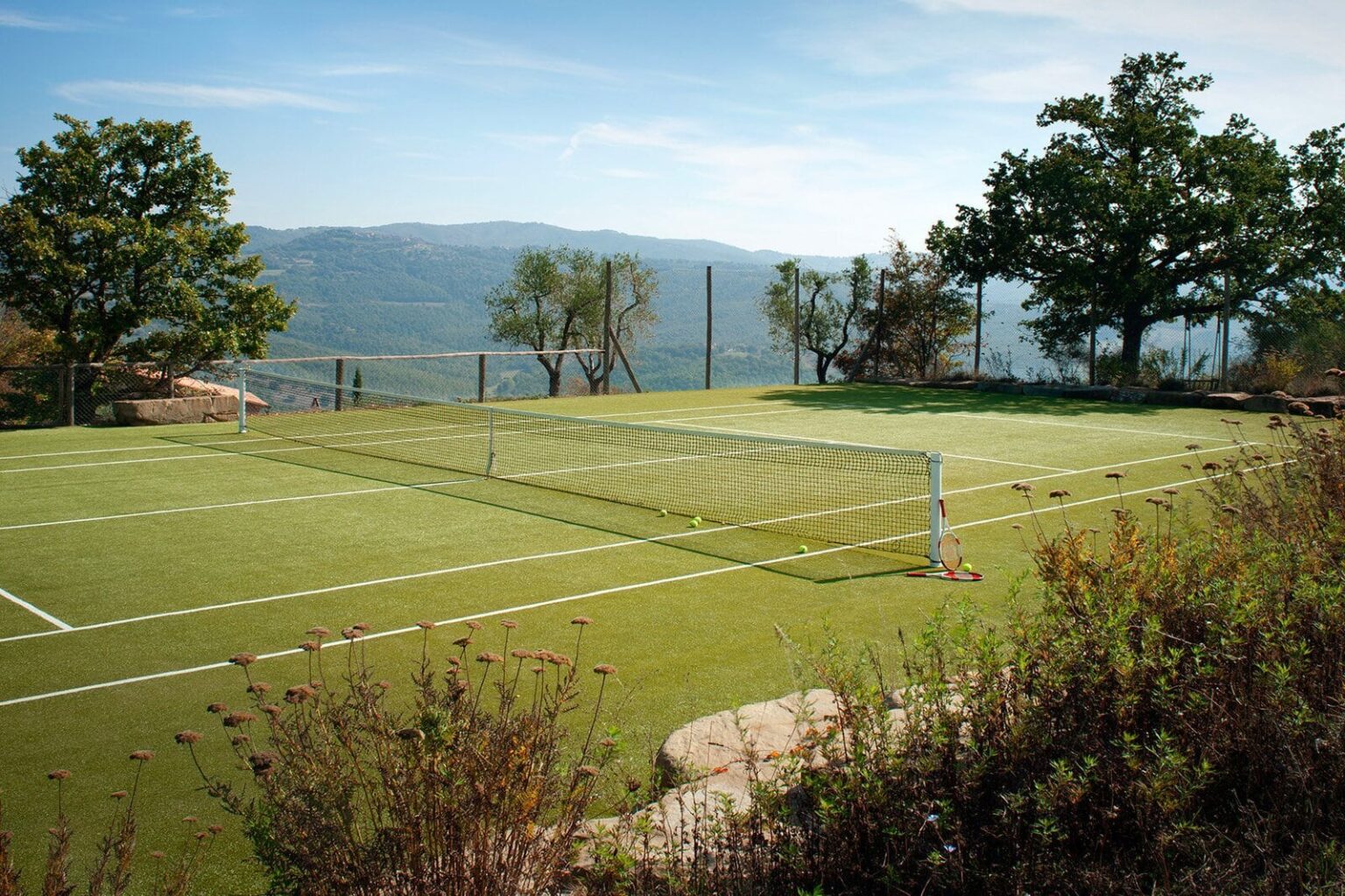 A scenic outdoor soccer field with a goal, surrounded by trees and mountains in the background, showcasing a clear blue sky and well-maintained artificial turf. | Only on SopranoVillas