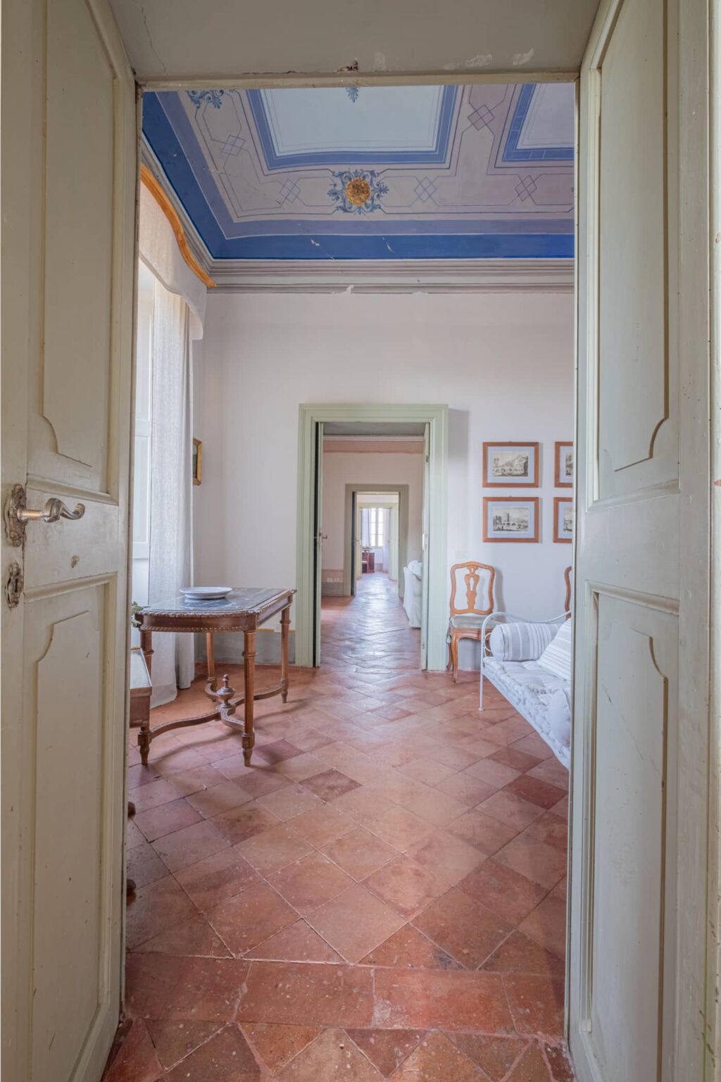 Interior view of a spacious, well-lit hallway featuring wooden doors, terracotta flooring, and framed artwork on the walls, leading to a cozy living area with a sofa and a table. | Only on SopranoVillas